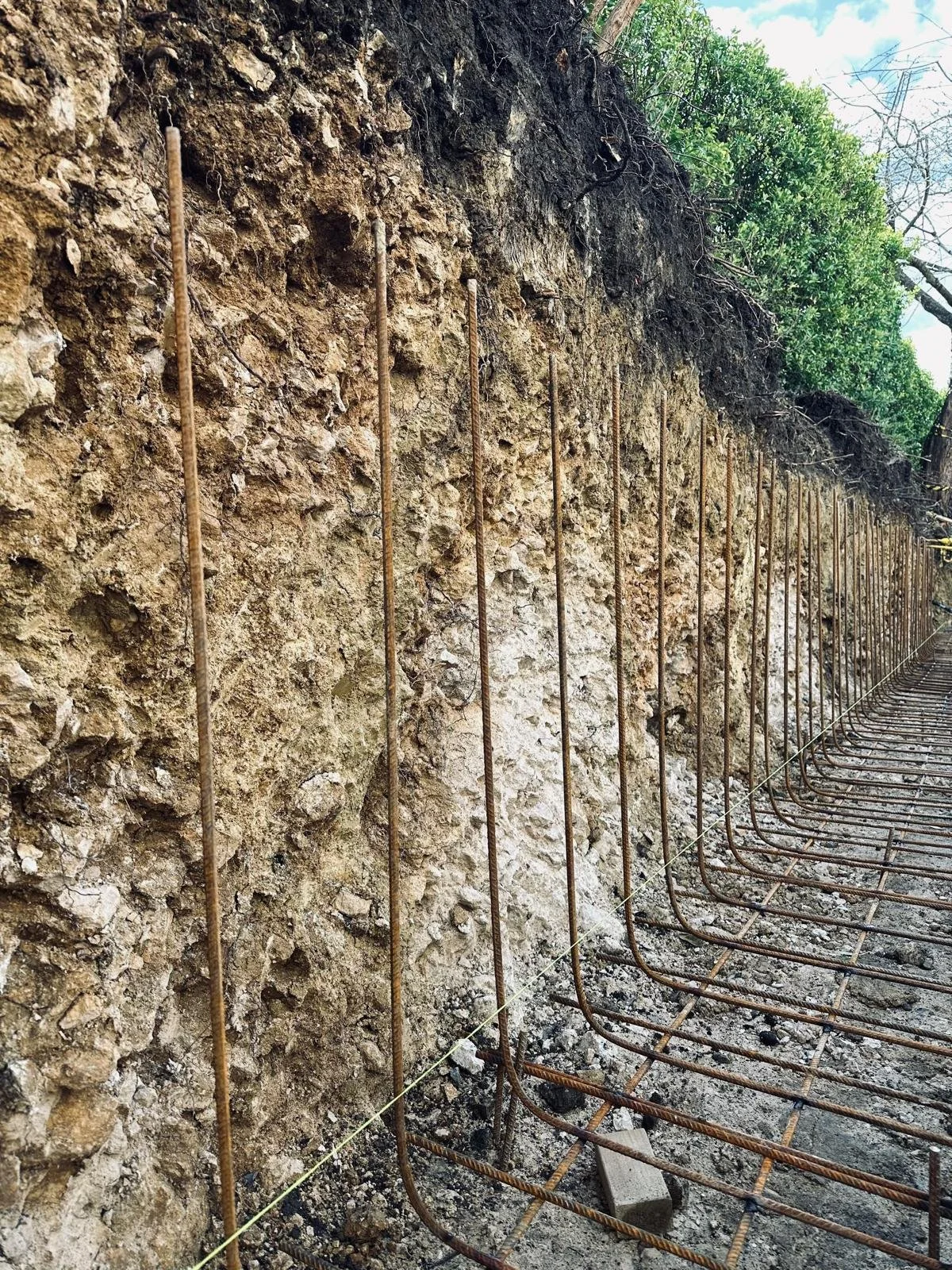 Vertical rebar rods and rebar mesh grid on concrete foundation next to a dirt wall, in a construction site.