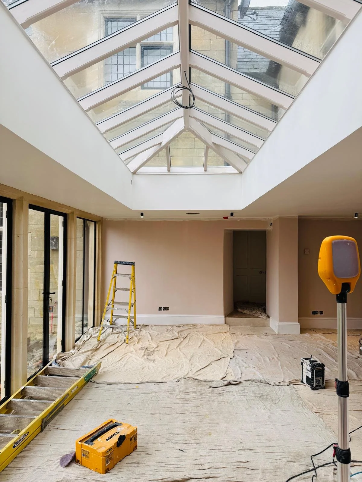 Interior of a house under renovation with a large glass ceiling, construction tools, a ladder, a portable work light, and covered flooring.