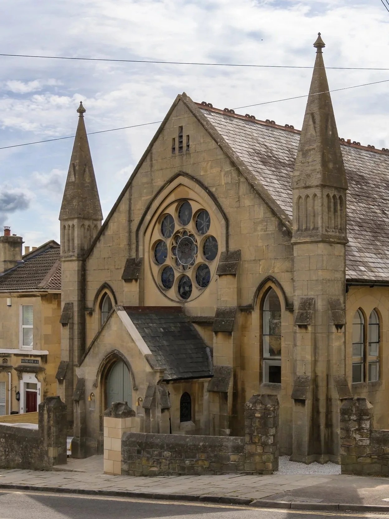 A stone church with Gothic architecture featuring a large circular stained-glass window and pointed spires, located in the city of Bath.
