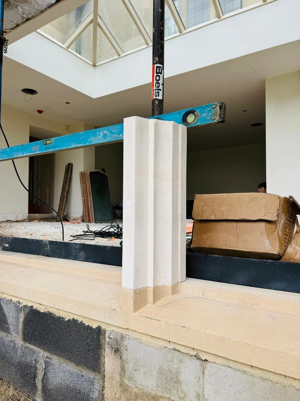 Construction site showing a concrete wall with insulation, a box, a level on a blue scaffolding, and construction materials inside a modern building with glass ceiling.