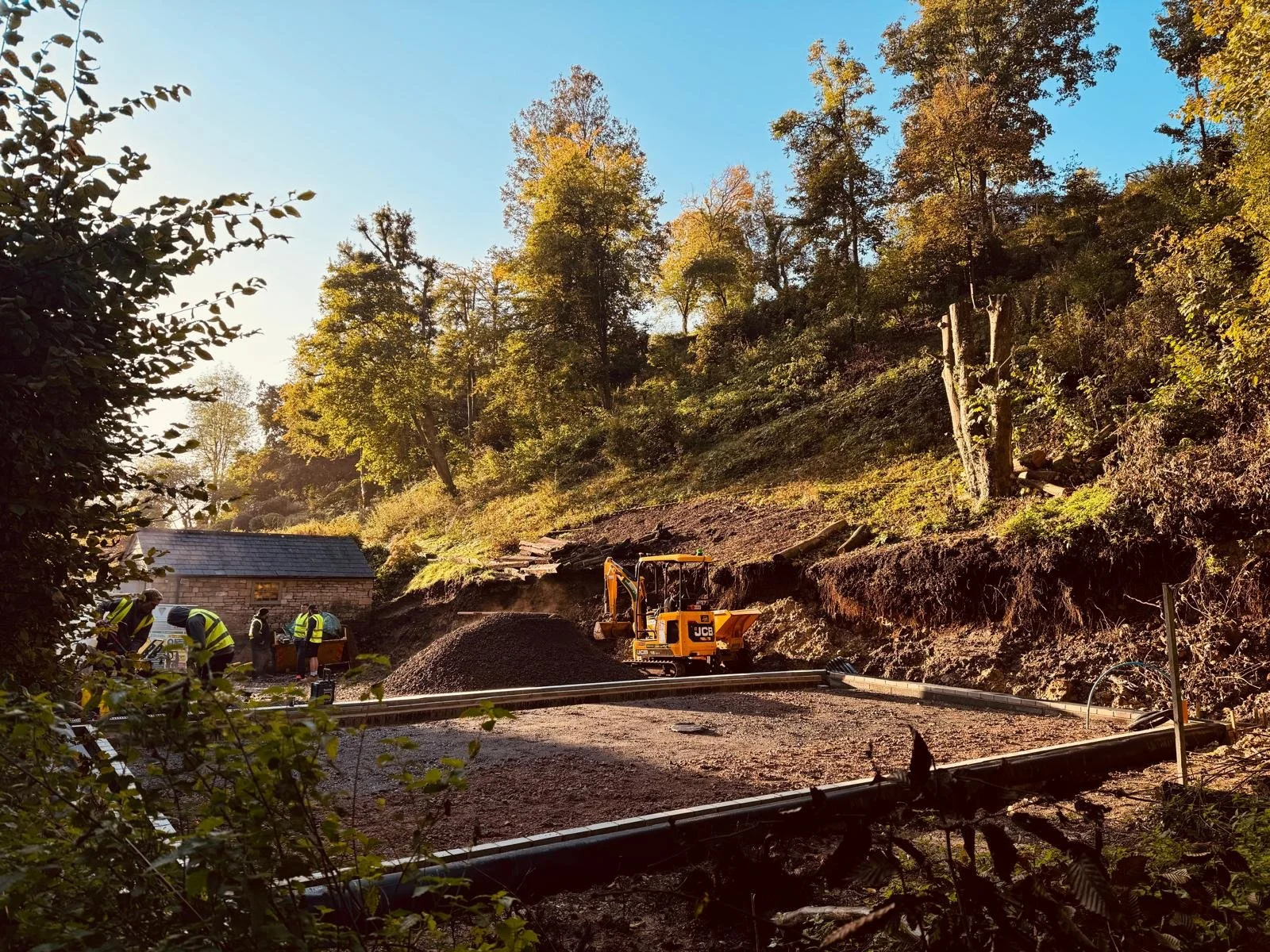 Workers at a construction site on a hillside using machinery and tools, with trees and a small building in the background under a clear blue sky.