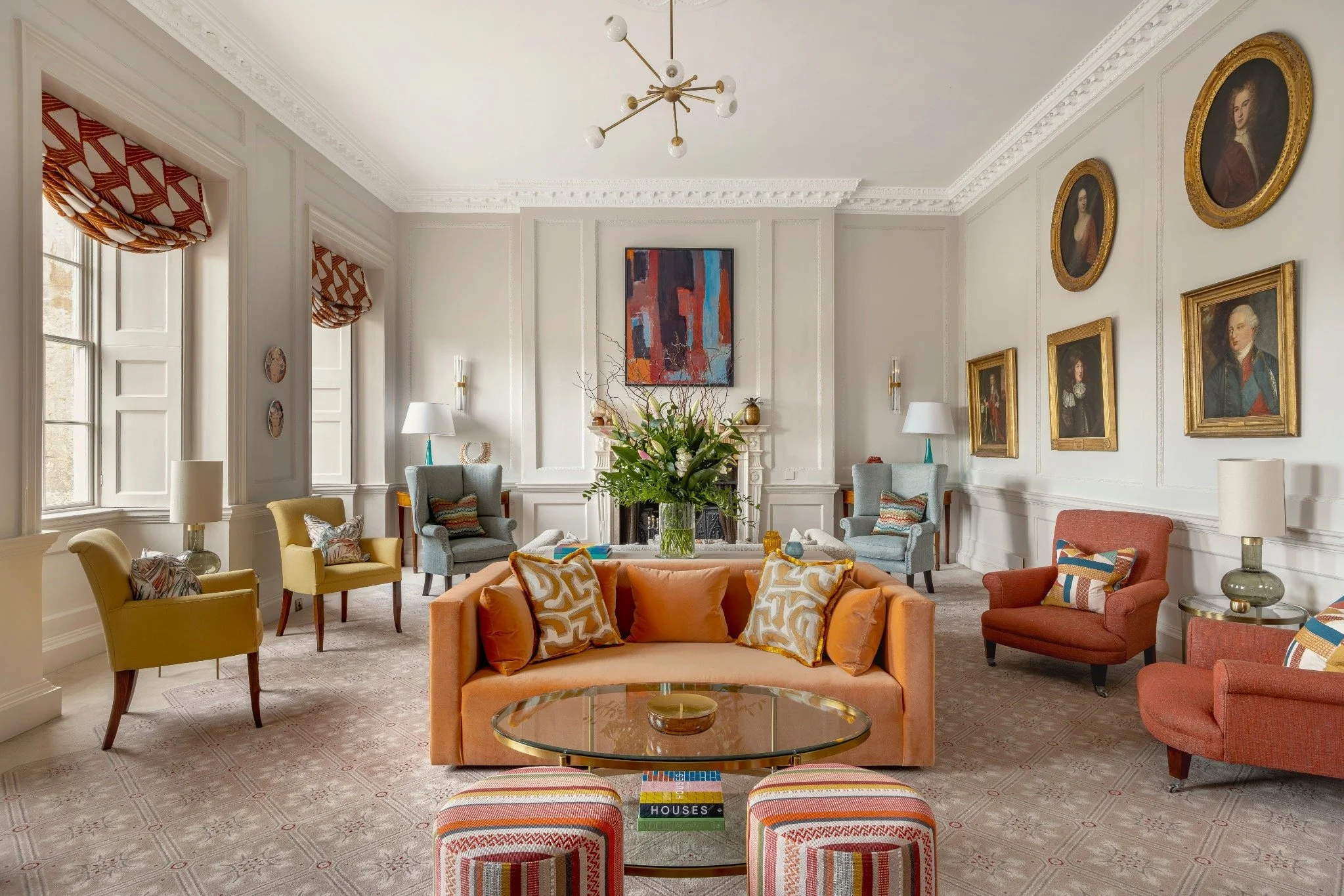 A spacious living room with white paneled walls and ornate crown molding, decorated with framed paintings, and featuring a variety of colorful chairs and sofas surrounding a glass coffee table with a flower arrangement.