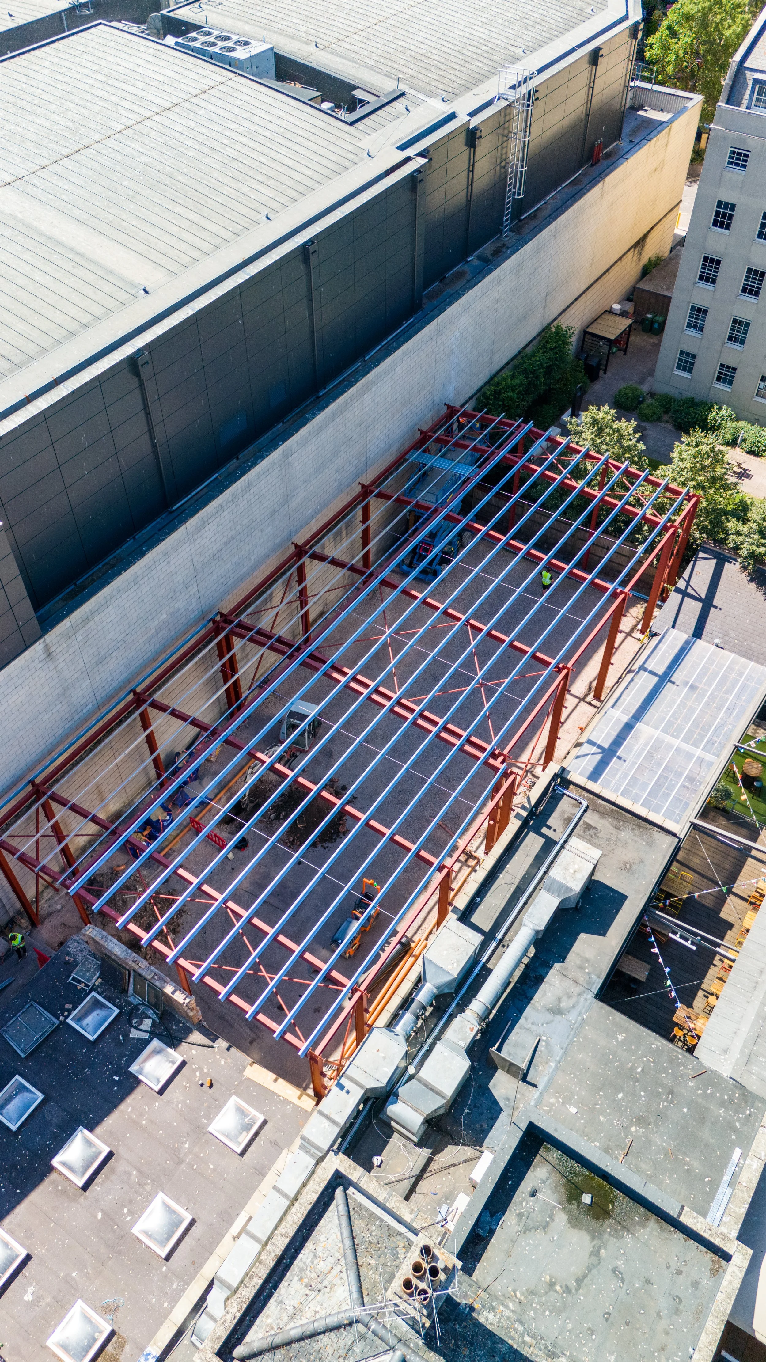 View of a building under construction with a metal framework on the roof and a black wall, surrounded by other buildings and trees, taken from a high angle.