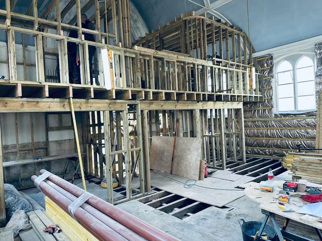 Interior of a building under construction with exposed wooden framing and scaffolding, along with windows on the right side and construction tools and materials scattered around.