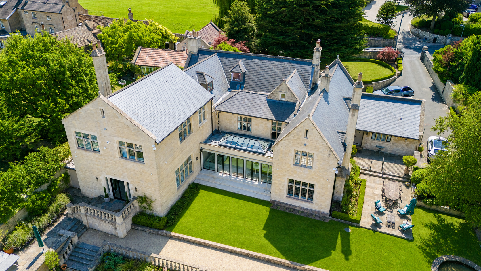Aerial view of a large, historic stone house with a steeply pitched roof, multiple chimneys, and large windows, surrounded by lush green trees and a well-maintained lawn, with a patio area featuring outdoor furniture.