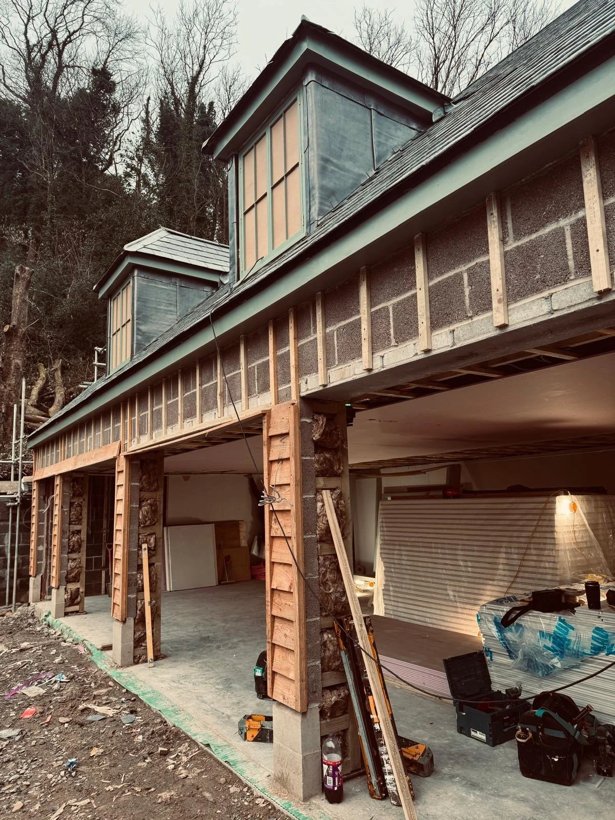 Construction site of a house extension with framing and an unfinished upper level, featuring two small attic windows and construction tools and materials around.