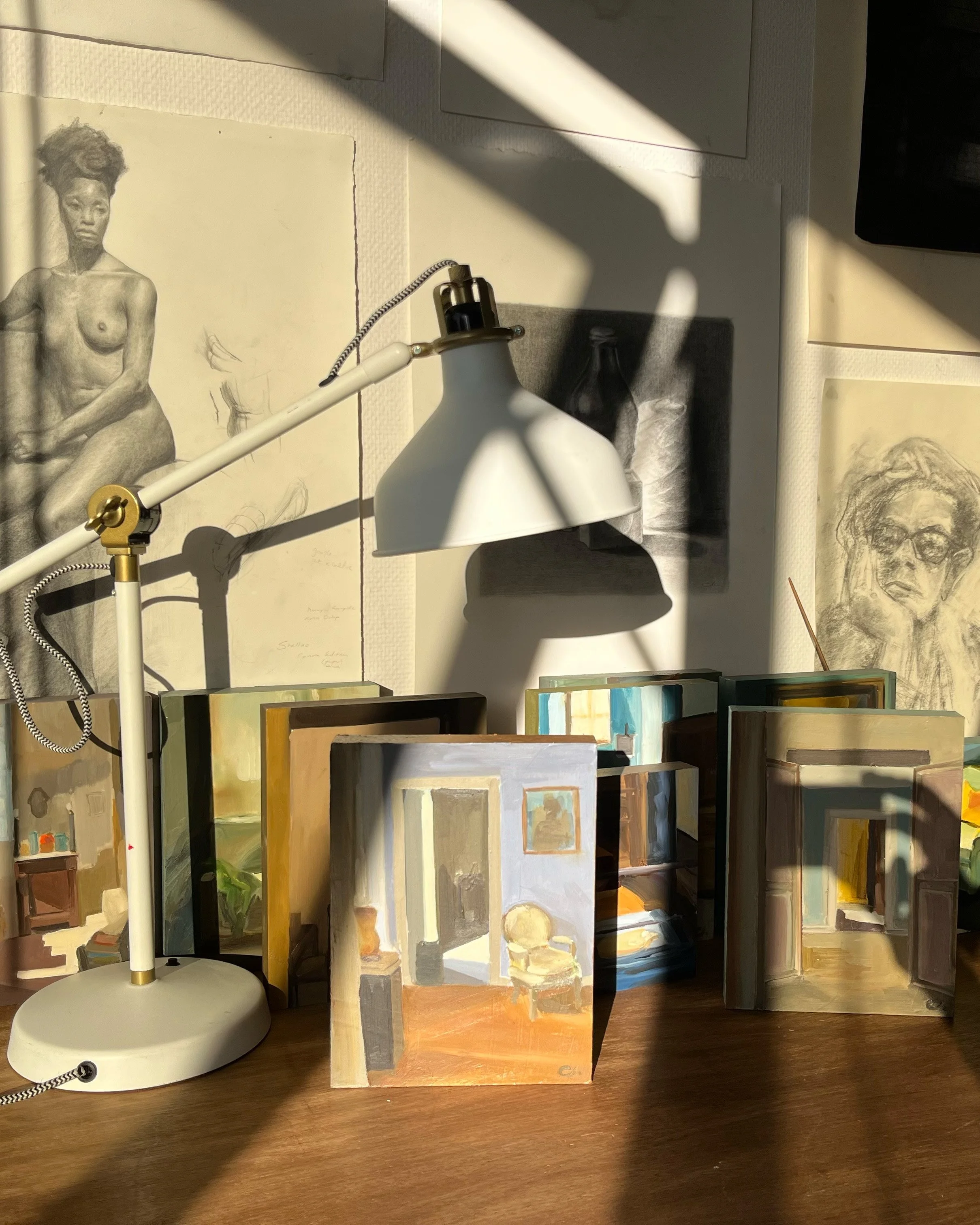 Une étagère avec plusieurs livres d'art et des dessins devant un mur blanc, éclairés par une lampe de bureau blanche. Des ombres de la lampe sont visibles sur le mur et les livres.