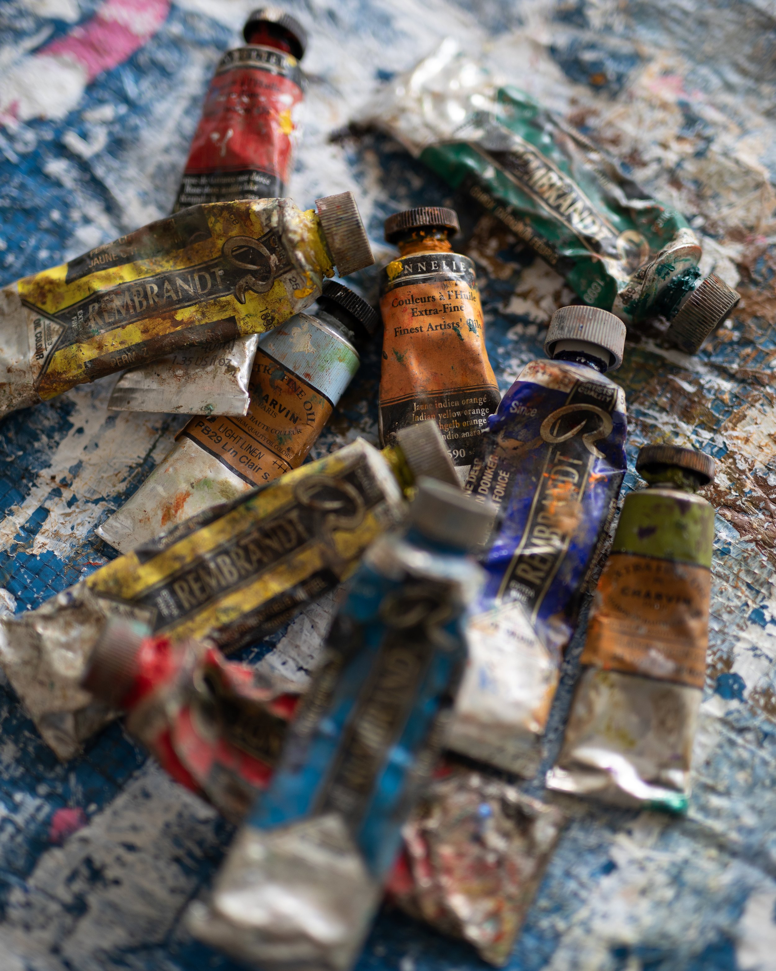 Tubes de peinture artistiques usagés, renversés et enroulés sur une surface peinte avec des taches de couleur, évoquant une scène de création artistique.