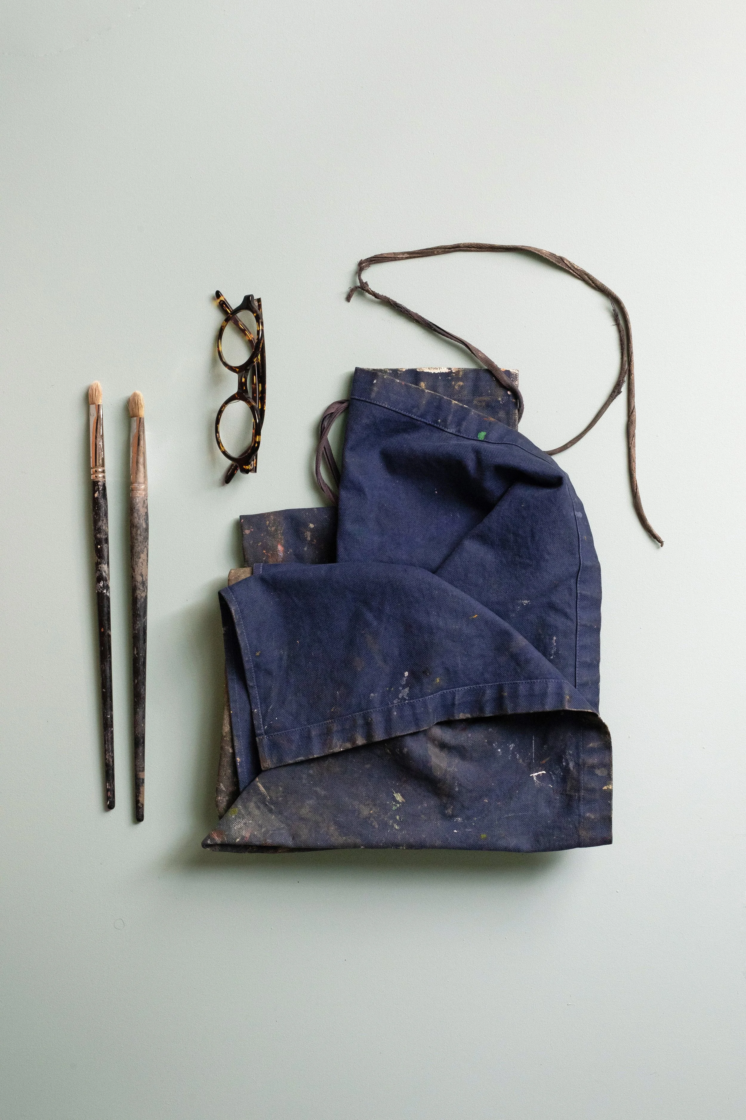 Deux pinceaux, une paire de lunettes, un tablier de peintre et un sac de peinture, tous couverts de peinture, disposés sur une surface claire.