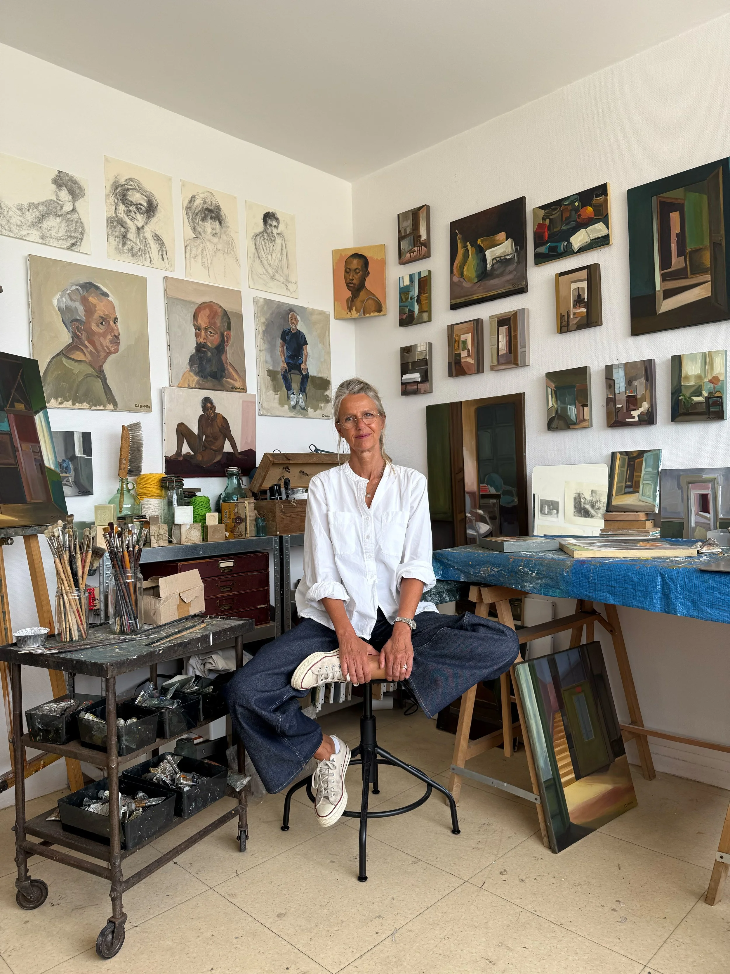 Une femme assise en pose méditative dans un atelier d'art, entourée de toiles et fournitures de peinture, avec des œuvres d'art sur les murs.
