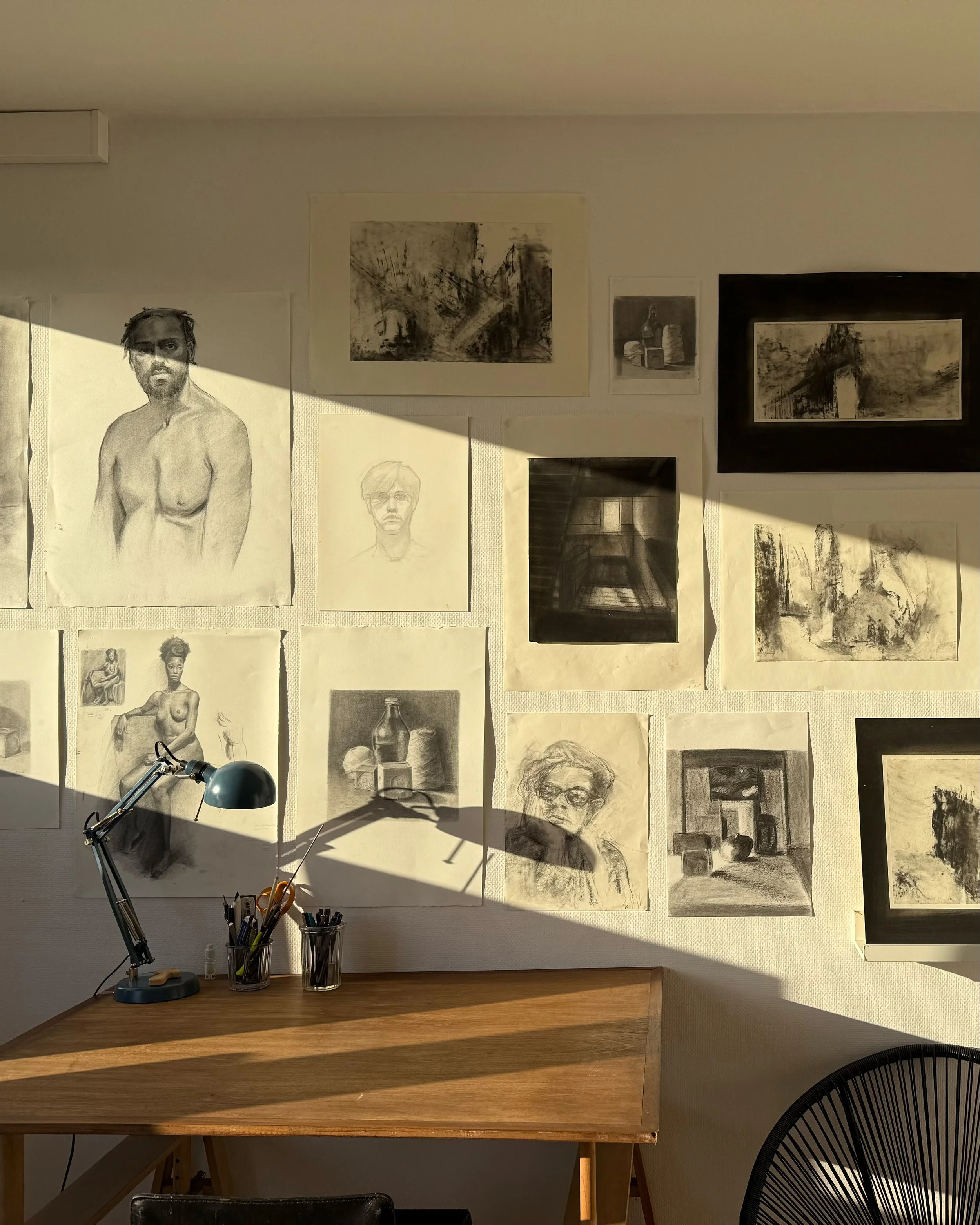 Mur blanc avec plusieurs dessins et peintures en noir et blanc, principalement de portraits et de paysages artistiques. Établi en bois avec une lampe de bureau et des contenants pour outils d'art. Ombres de lumière naturelle projetant une ambiance créative.