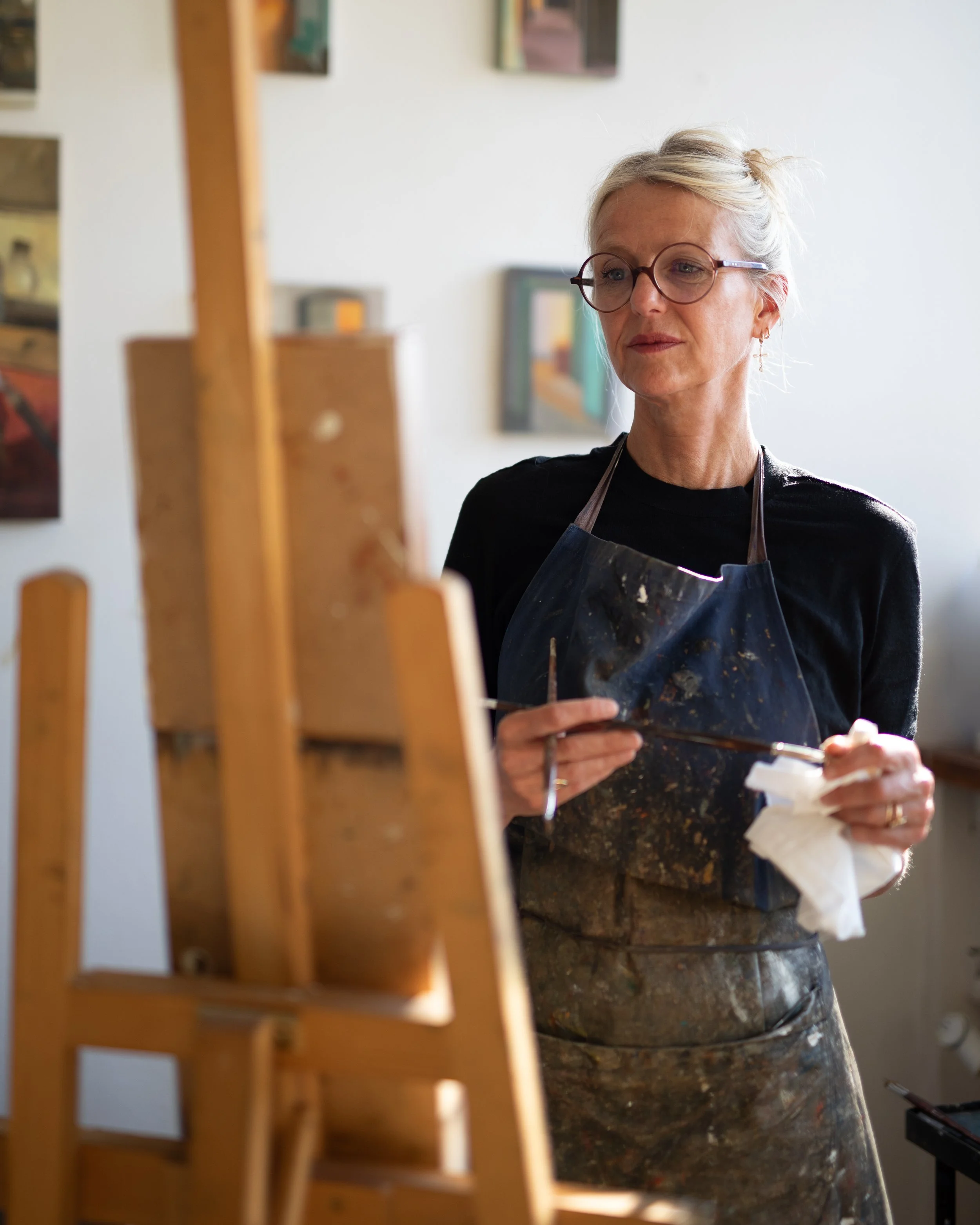 Une femme blonde portant des lunettes rondes et un tablier sale, probablement en train de peindre ou de réaliser une œuvre artistique dans un espace intérieur, avec des peintures accrochées au mur en arrière-plan.