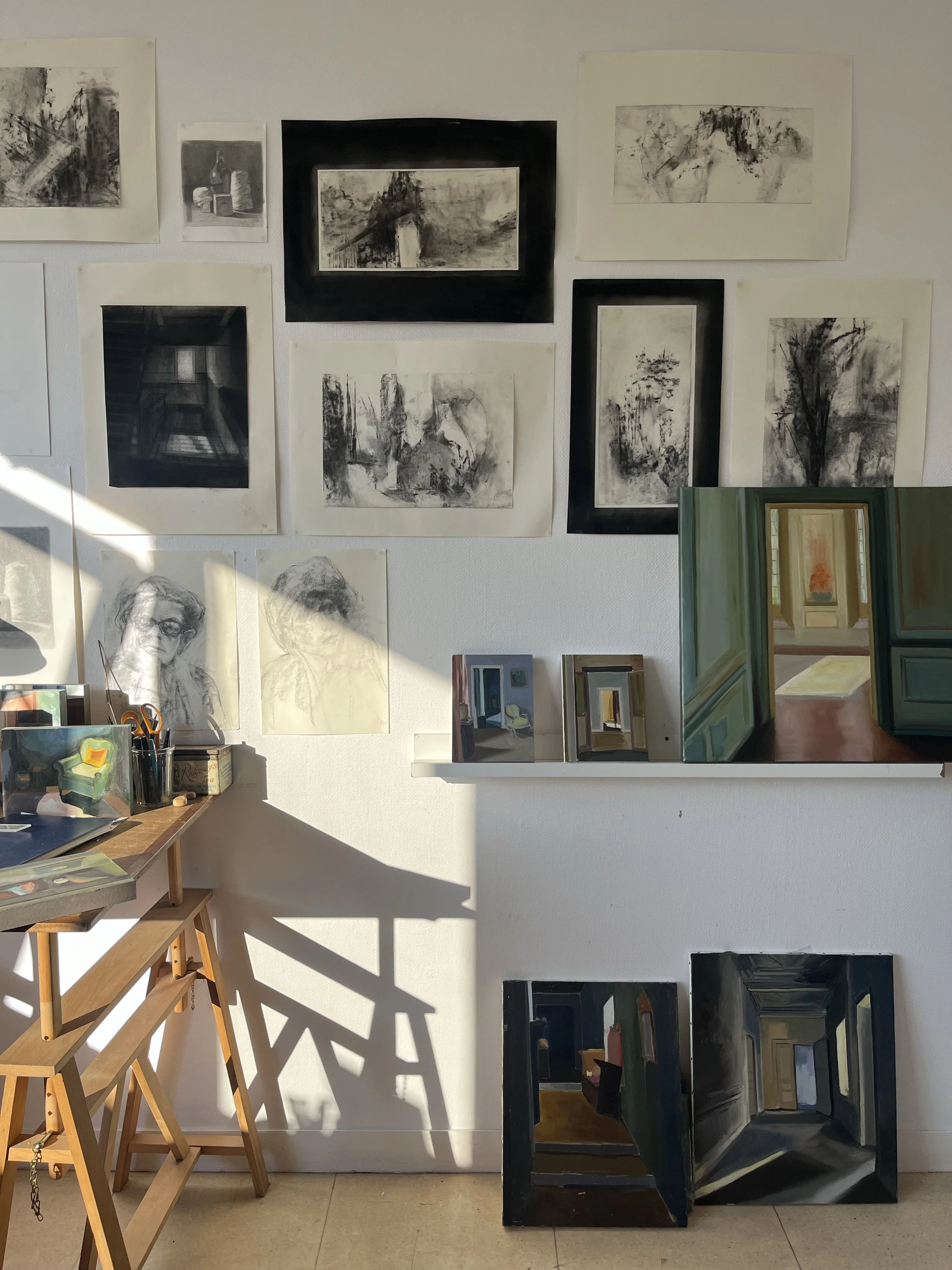 Une scène dans un atelier d'art avec plusieurs œuvres en noir et blanc accrochées au mur. Il y a un échafaudage en bois avec des outils à côté et plusieurs toiles. La lumière du soleil crée des ombres sur le mur.