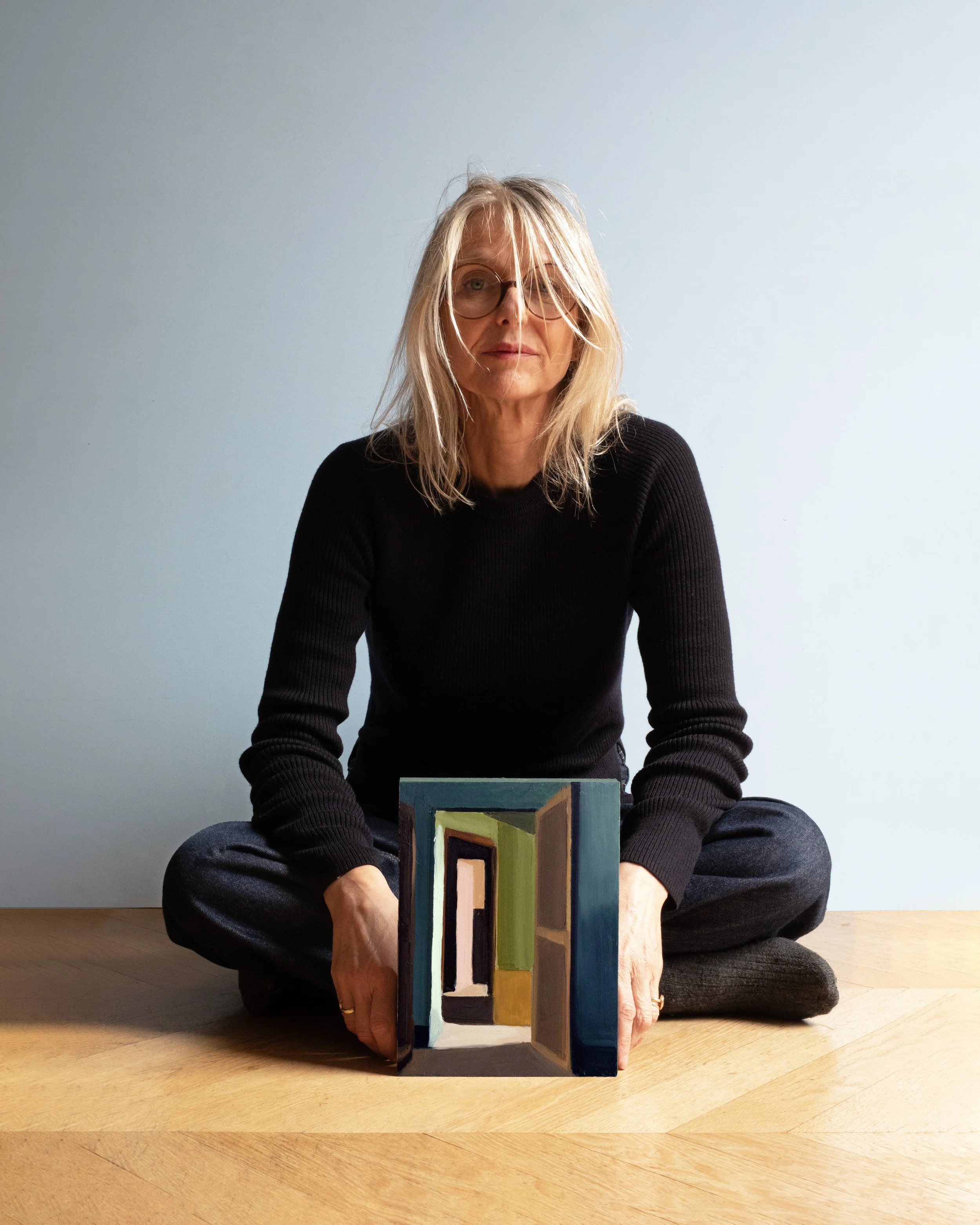 A middle-aged woman with blonde hair and glasses, sitting cross-legged on the floor, holding a colorful abstract painting in front of her. She is wearing a black sweater and jeans, with a neutral facial expression. The background is a plain, light-colored wall.