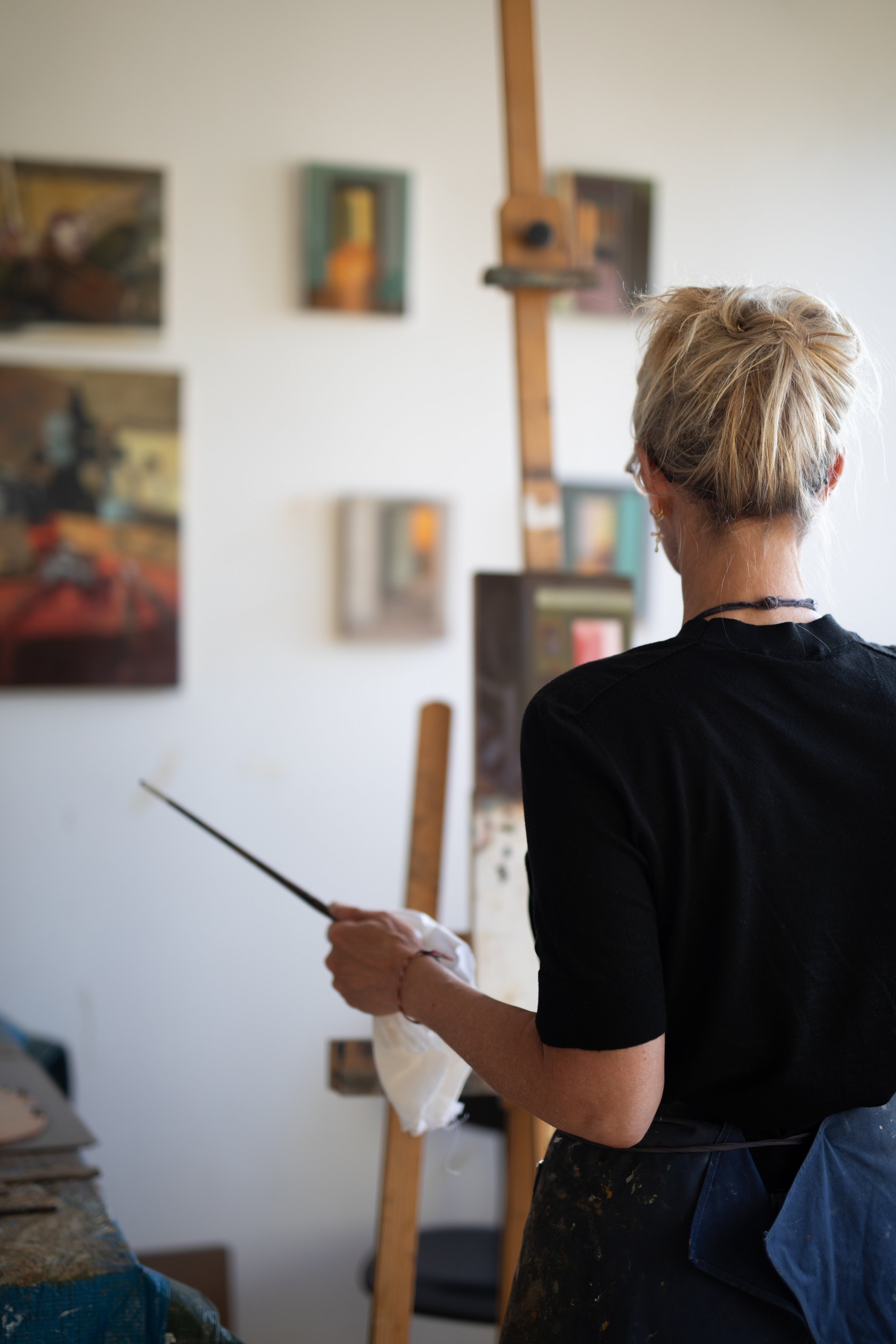 Une femme avec des cheveux blonds en chignon, portant un sweat noir, regarde une toile dans un atelier d'art, tenant un pinceau et un chiffon blanc.