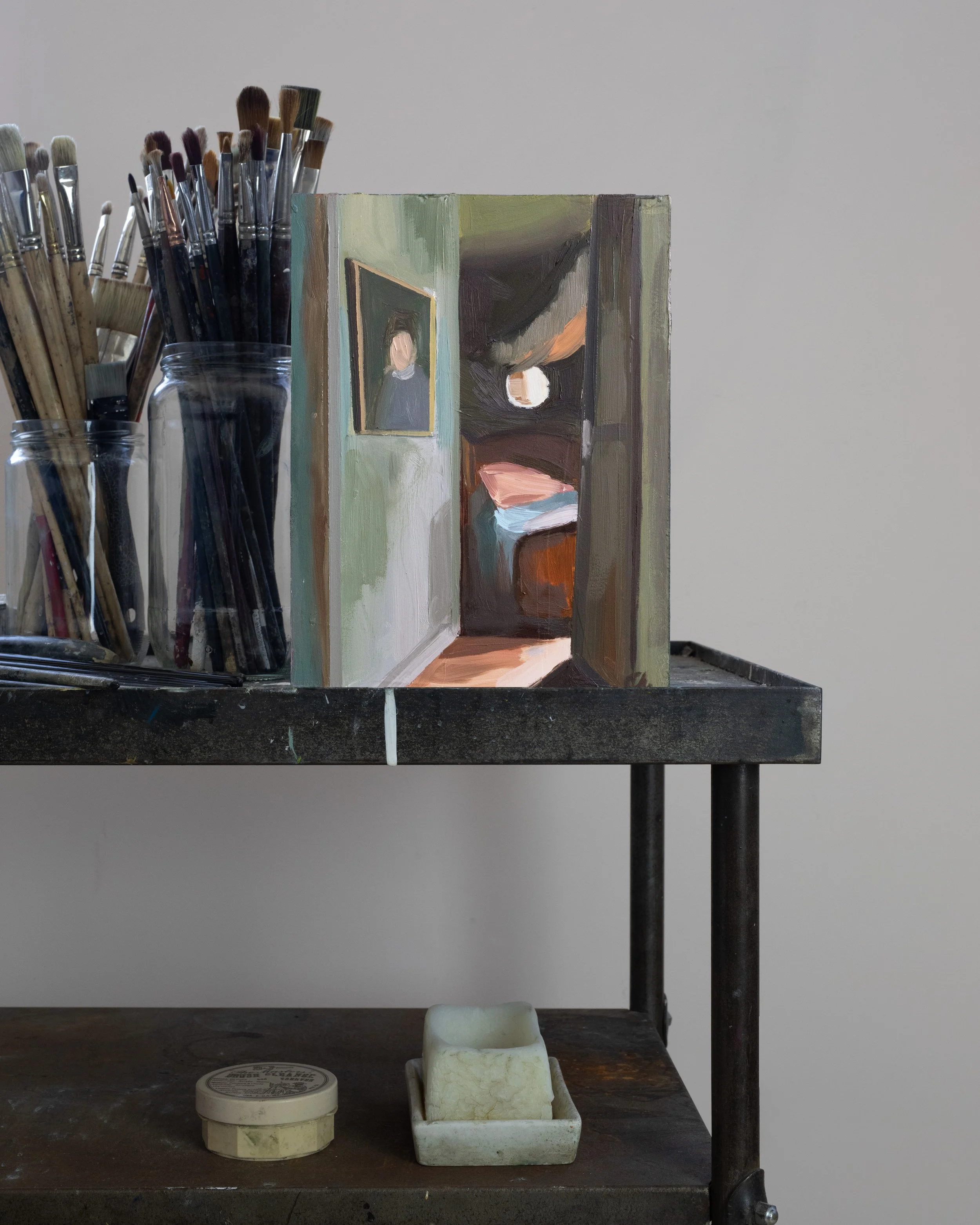 Une table de travail d'artiste avec un ensemble de pinceaux dans des bocaux en verre à gauche, une maquette ou un painting en cours représentant l'intérieur d'une pièce avec un portrait au mur, et plusieurs objets de sculpture ou des matériaux en bas, sur la surface de la table. Le fond est une surface murale simple de couleur claire.