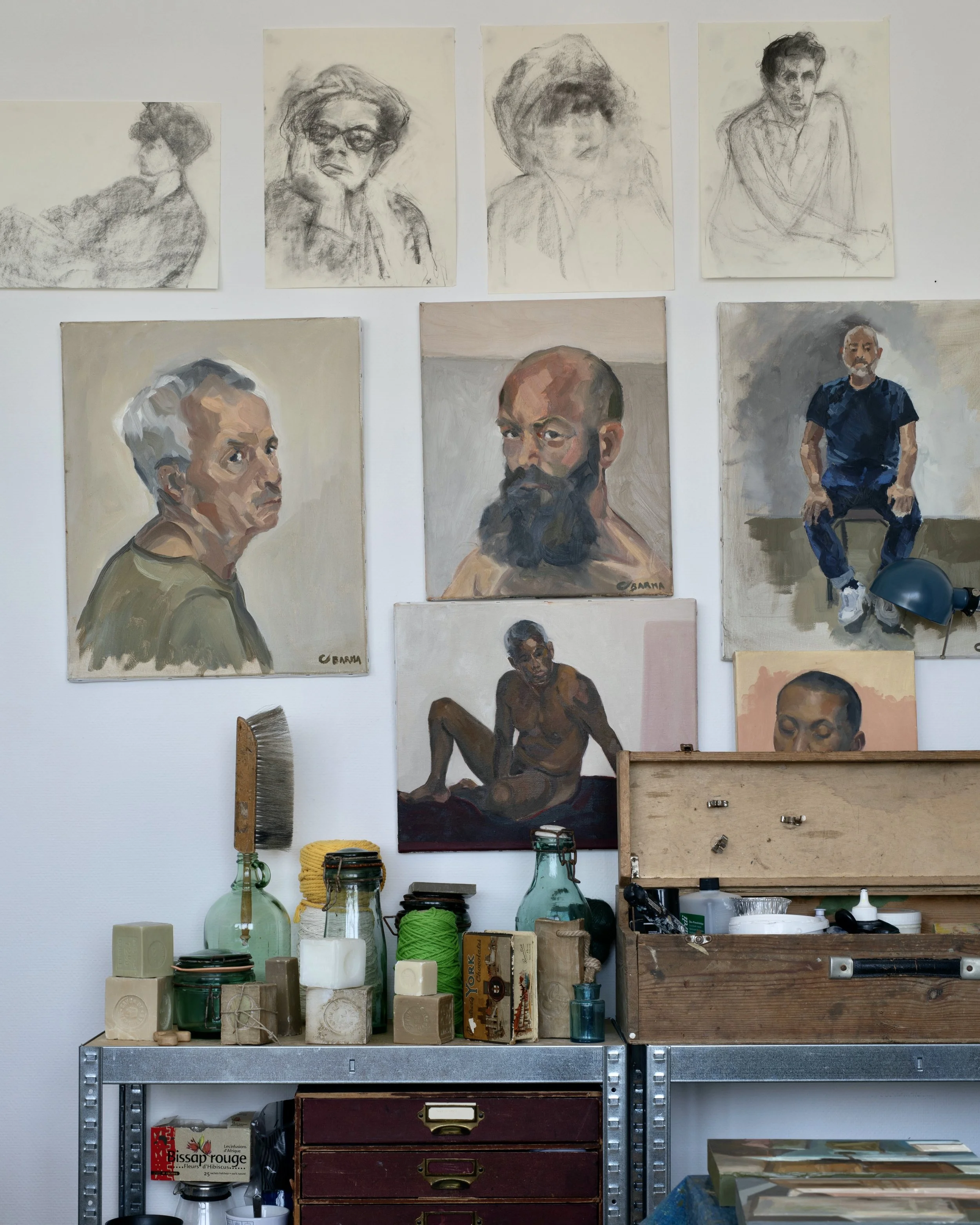 Une étagère contenant des bocaux en verre, des livres, des barils en bois, un tableau de portraits à l'huile, un ventouse, un pinceau, une caisse en bois avec des petits objets, un tableau avec une personne en train de dessiner, et d'autres objets.