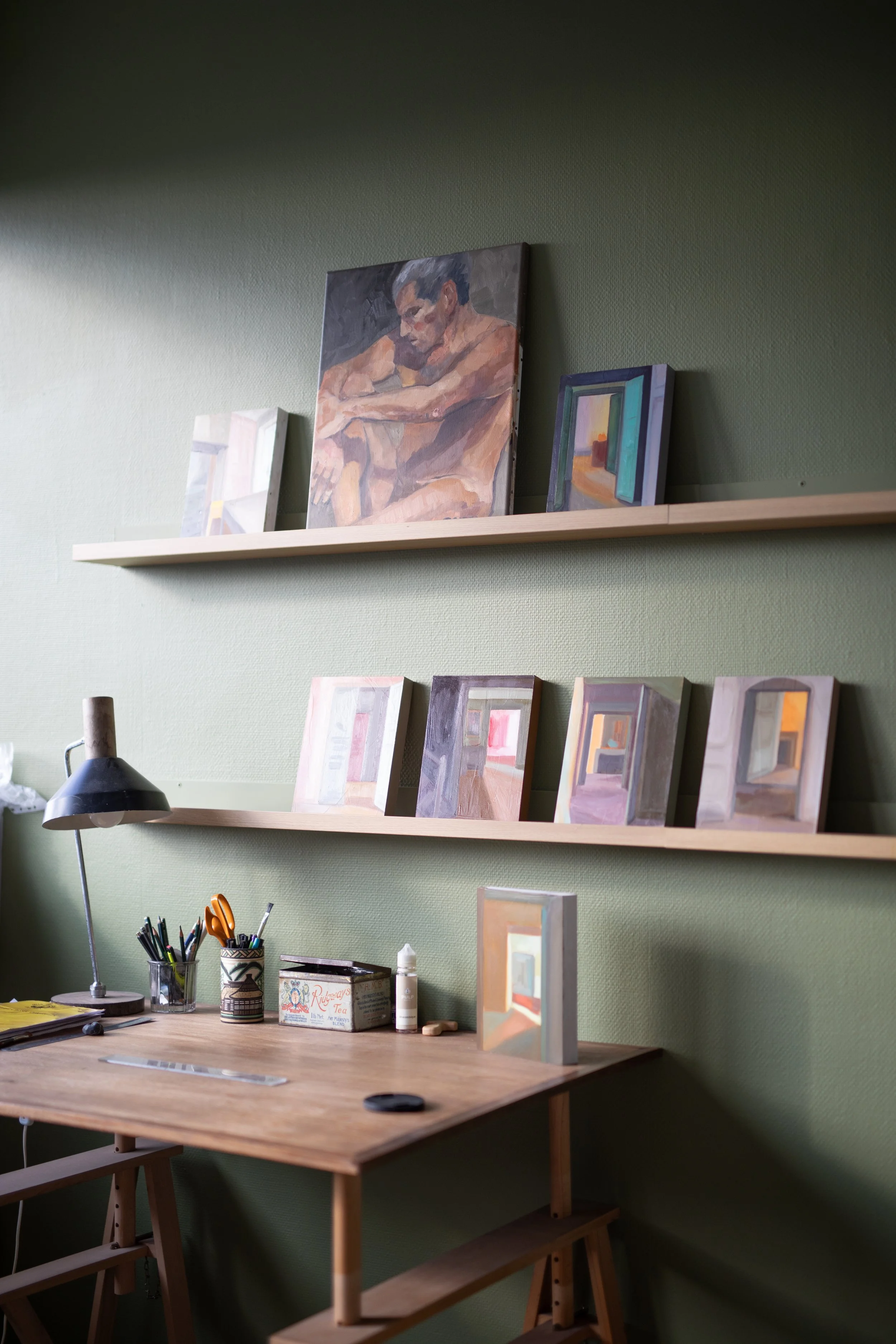 Un espace de travail avec un bureau en bois, un lampadaire noir, des pinceaux, un pot de peinture, une boîte de thé et des peintures exposées sur deux étagères vertes. Plusieurs peintures de style différent sont visibles.