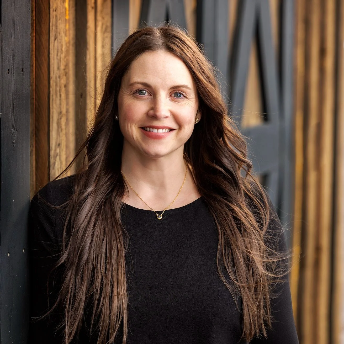 A woman with long brown hair wearing a black top, standing in front of a rustic wooden background, smiling at the camera.