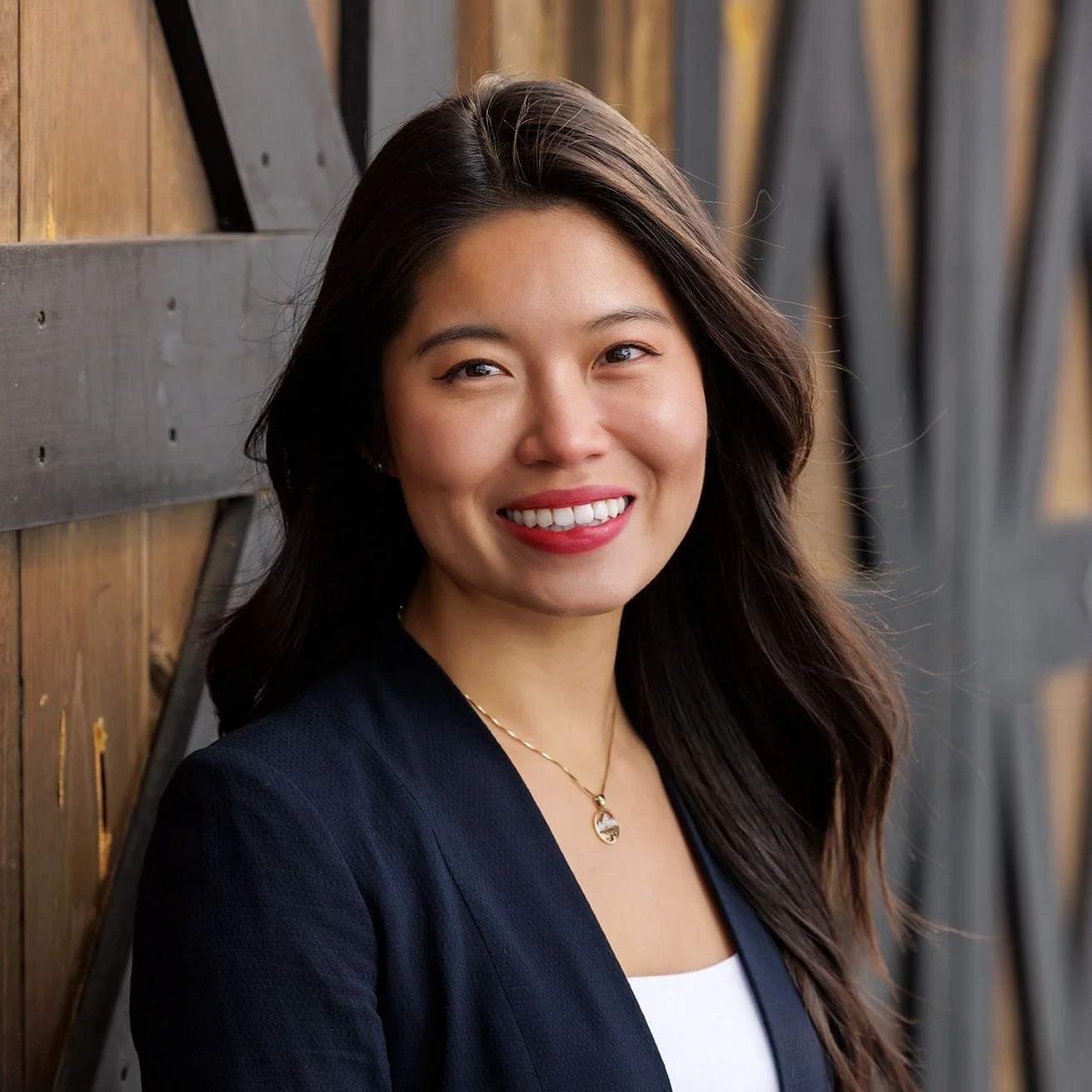 A smiling Asian woman with long dark hair, wearing a dark blazer, a white top, and a gold necklace, standing against a wooden and metal background.