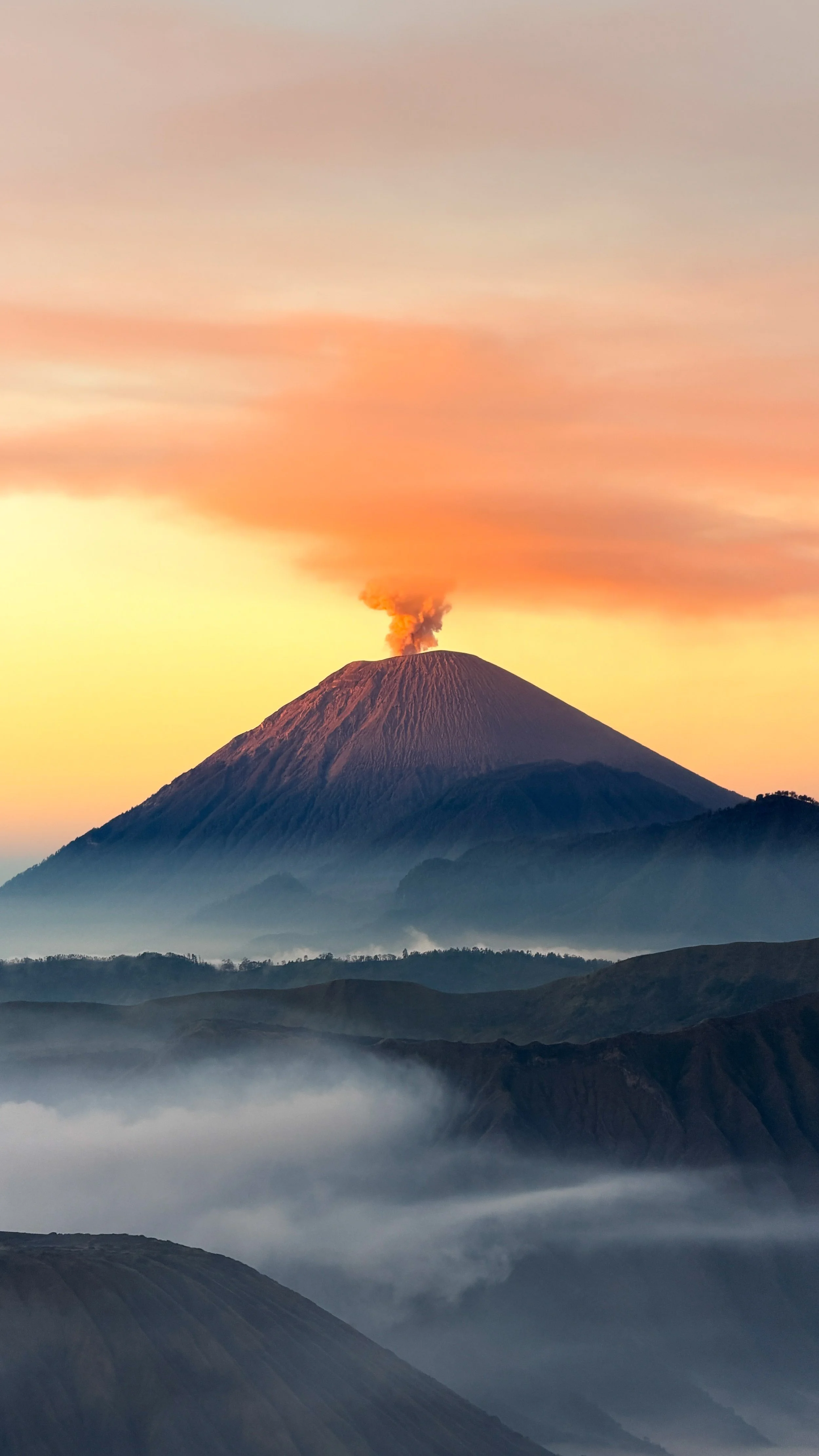 Volcano erupting with ash and smoke at sunrise, with multiple mountain ridges and low clouds in the foreground. Photo taken by Hailey Tasker, a registered therapist in Ontario, offering remote counselling for young adults