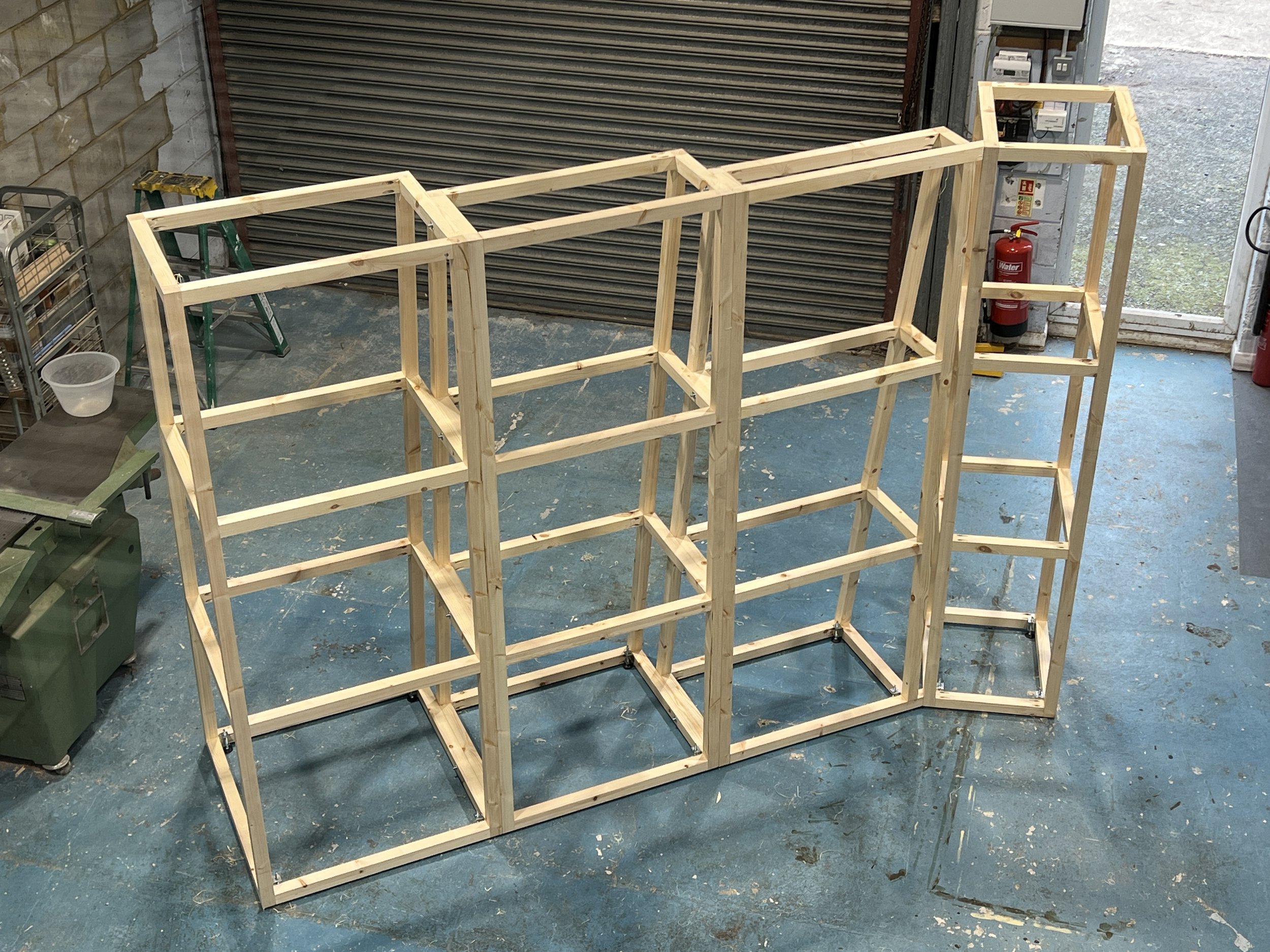 Wooden frame structure under construction in a workshop, surrounded by tools and equipment.