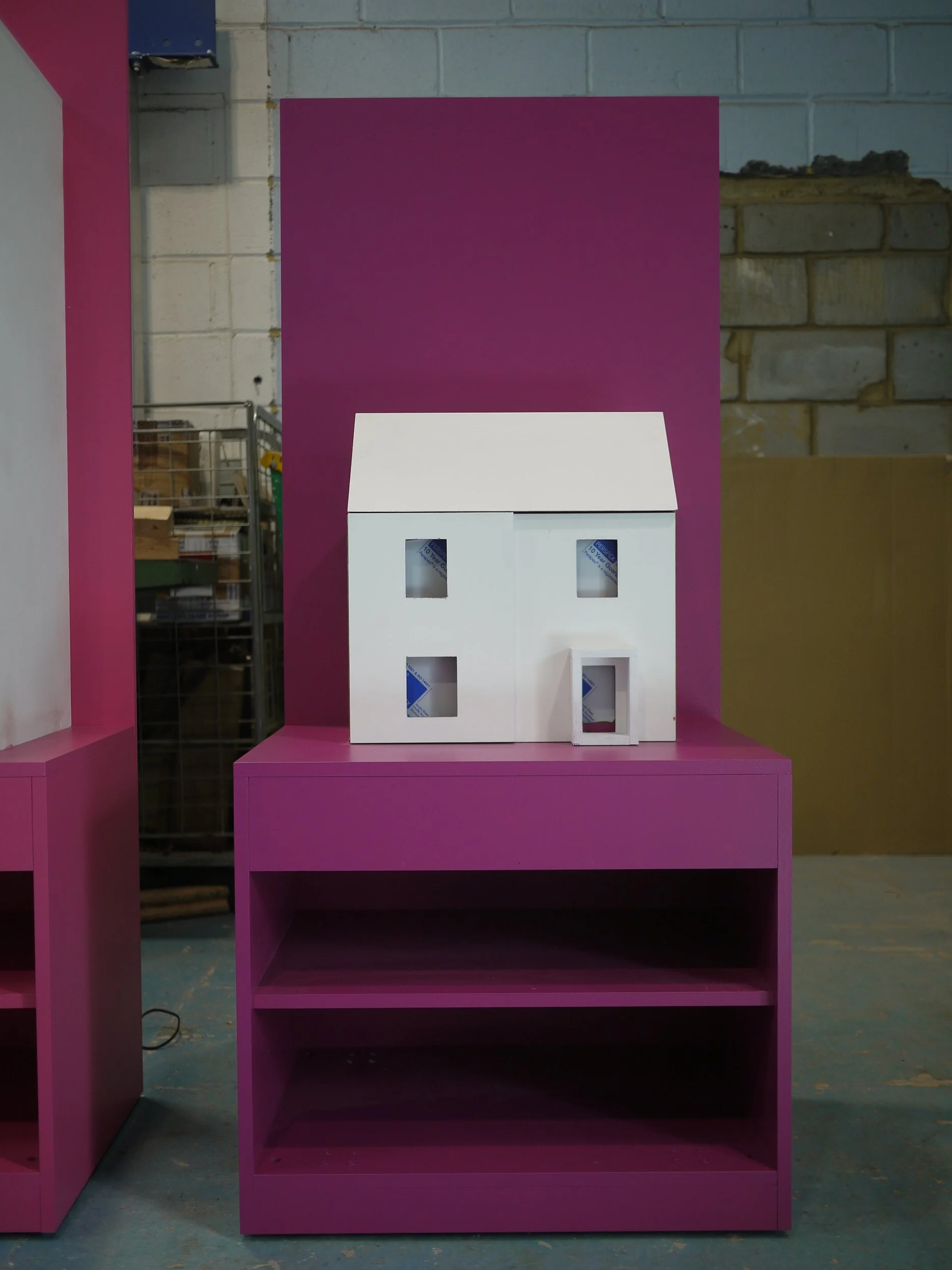 White cardboard house with windows and door, placed on a pink display stand in front of a purple backdrop, in a warehouse or workshop setting.