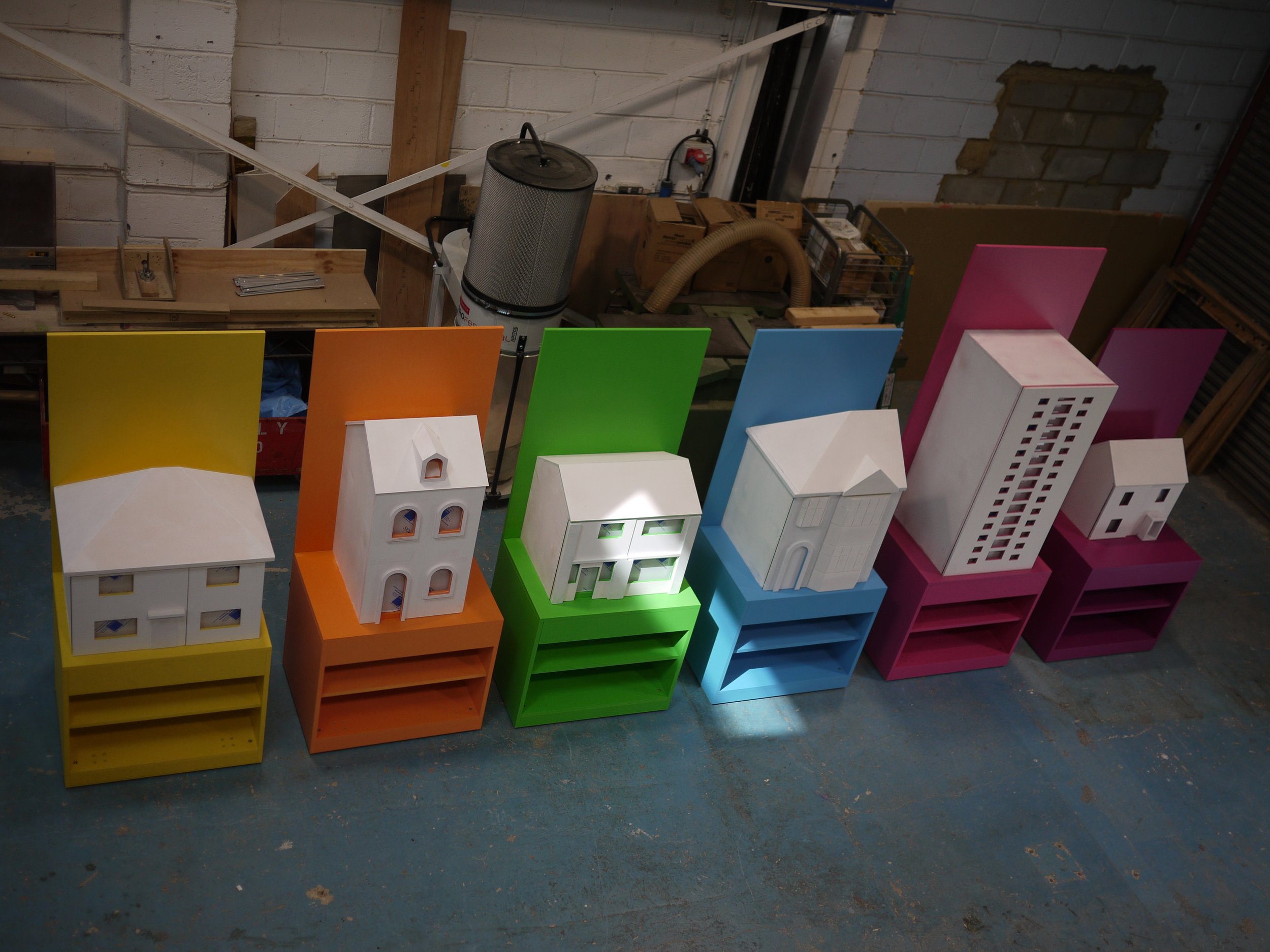 Colorful miniature building models displayed in a row, including houses and high-rise structures, placed on top of colorful shelves in a workshop or store setting with a concrete wall background.