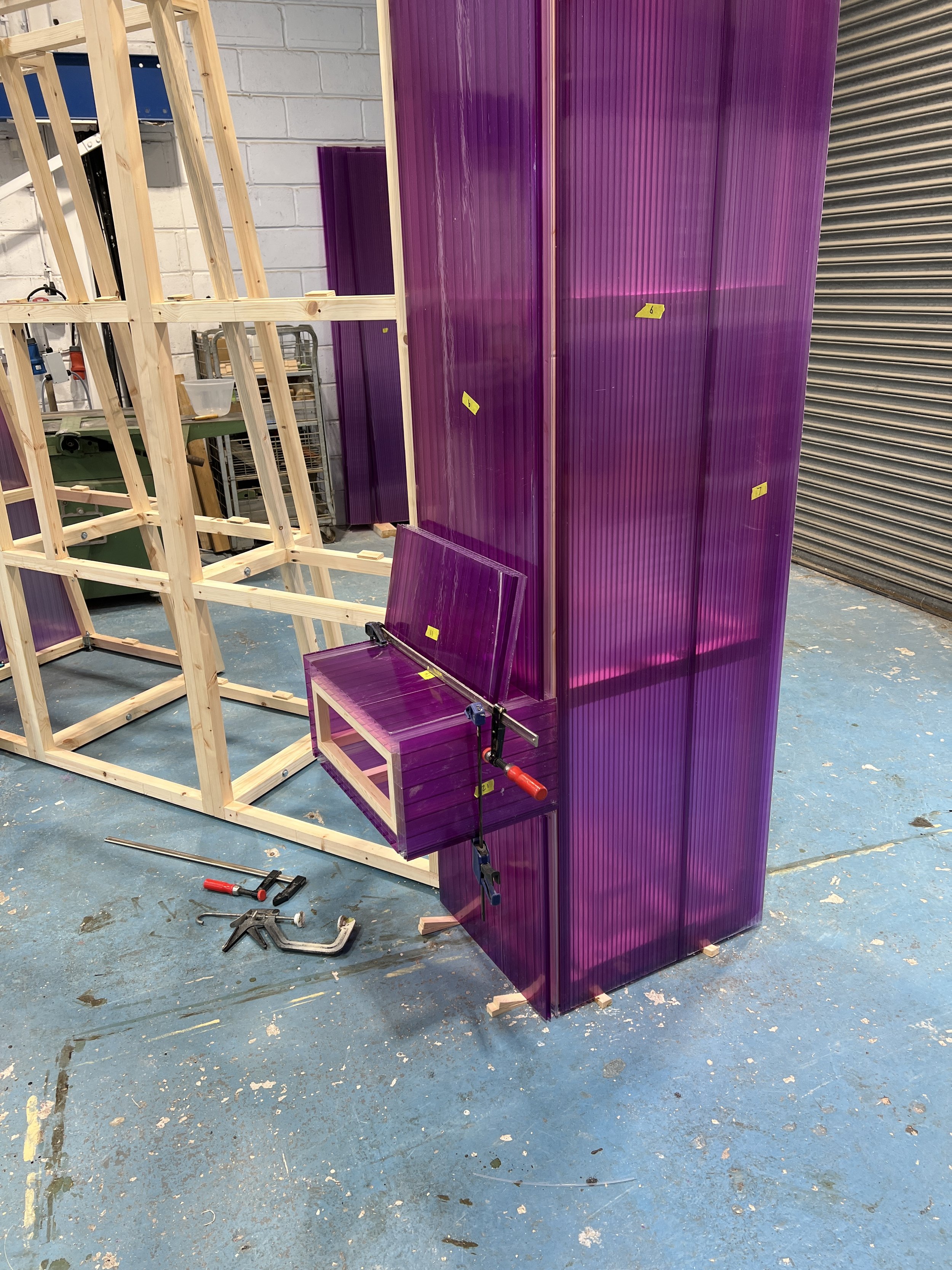 Workshop scene with a wooden frame structure and purple acrylic panels, some clamped to the frame, on a blue workbench with tools and clamps scattered on the floor.