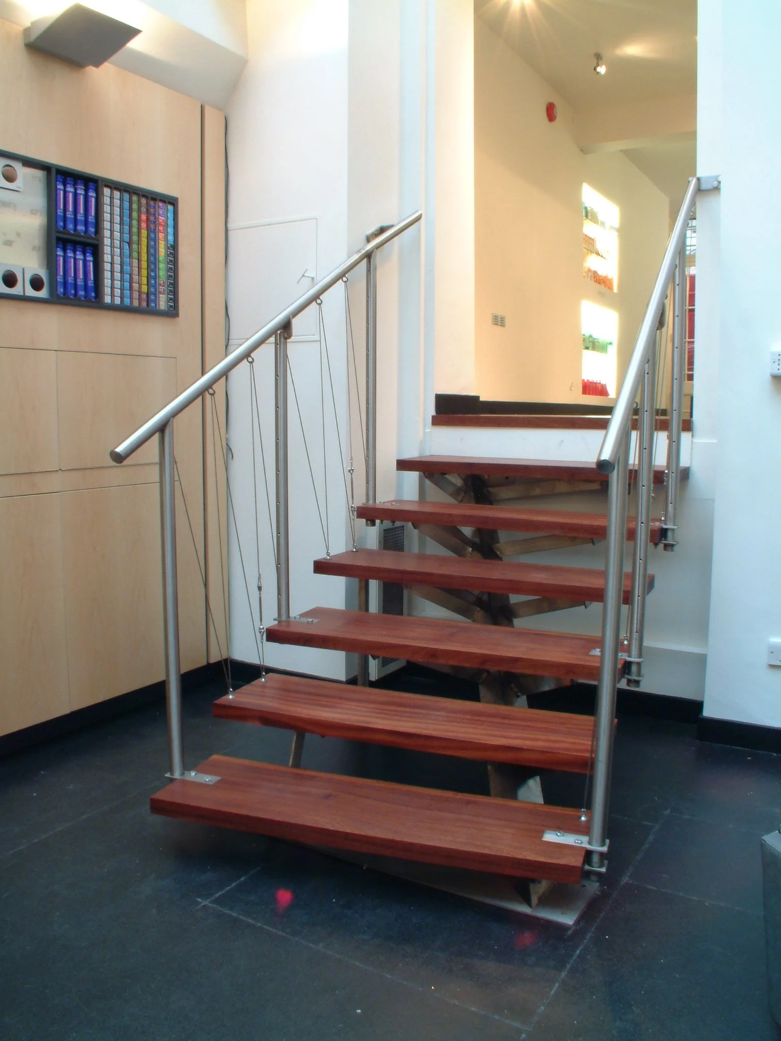 Interior of a modern building with a spiral staircase featuring wooden steps and metal railings. The background shows a wall with colorful lighted display shelves, and the flooring is dark.