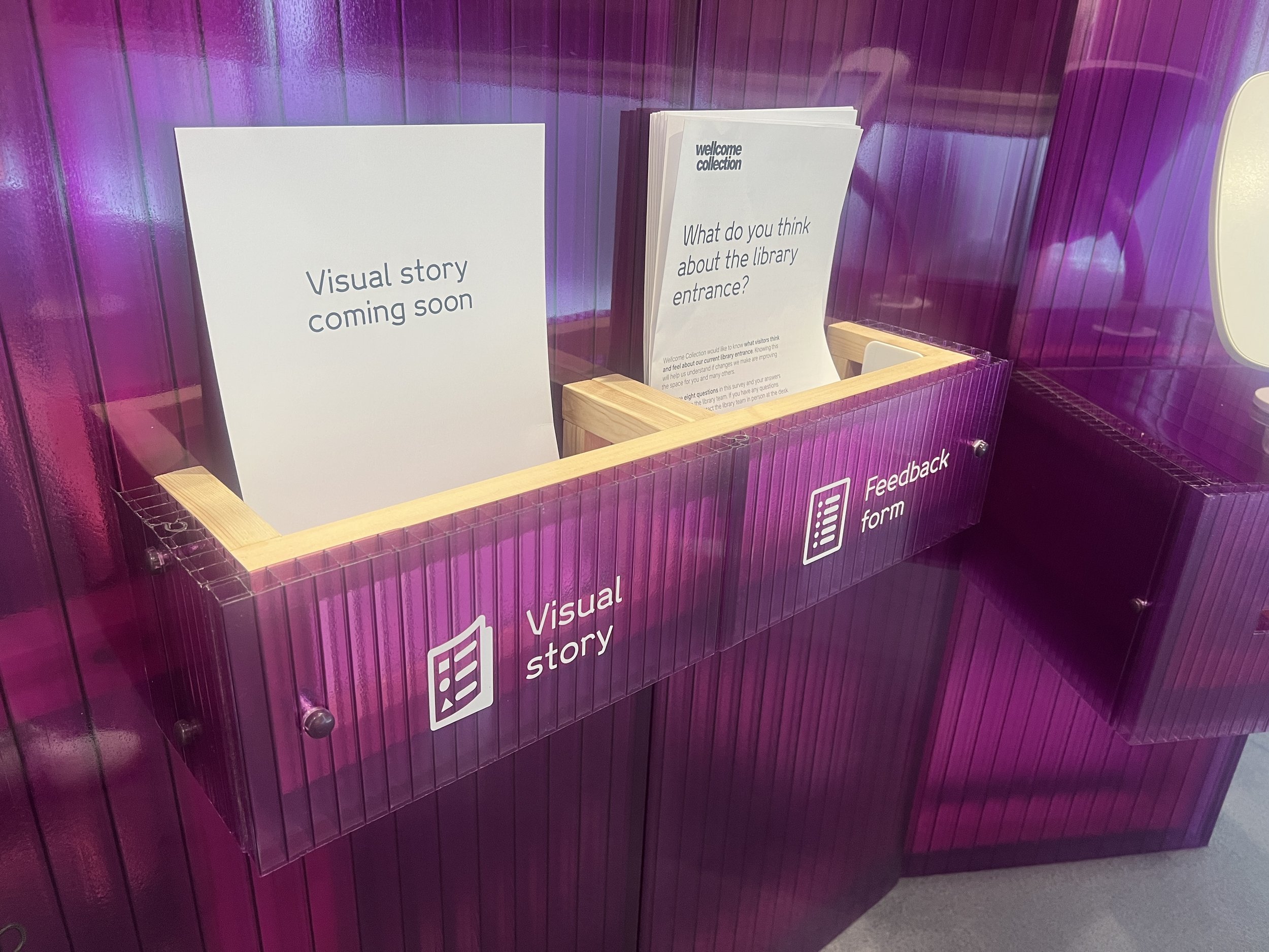 A purple library display with signs that read 'Visual story coming soon' and 'What do you think about the library entrance?'. There is a feedback form icon on the side.