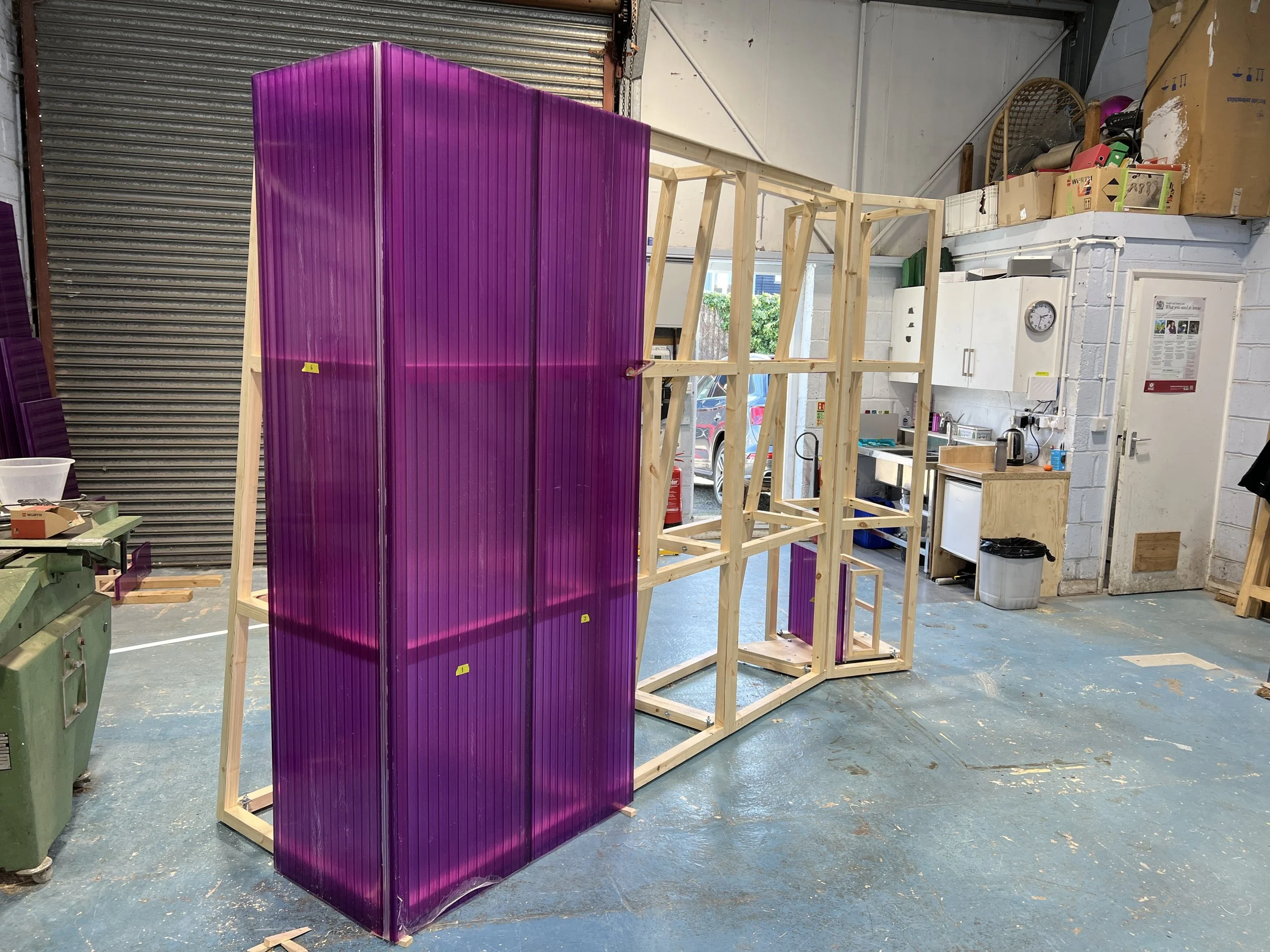 A workshop with a large purple translucent panel leaning against a wooden frame structure. The workshop has a garage door, workbench, and supplies, with a window and some greenery outside.