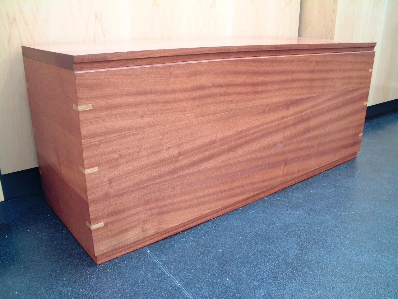 A rectangular wooden storage bench with a flat top, placed against a light wooden wall on a dark gray floor.