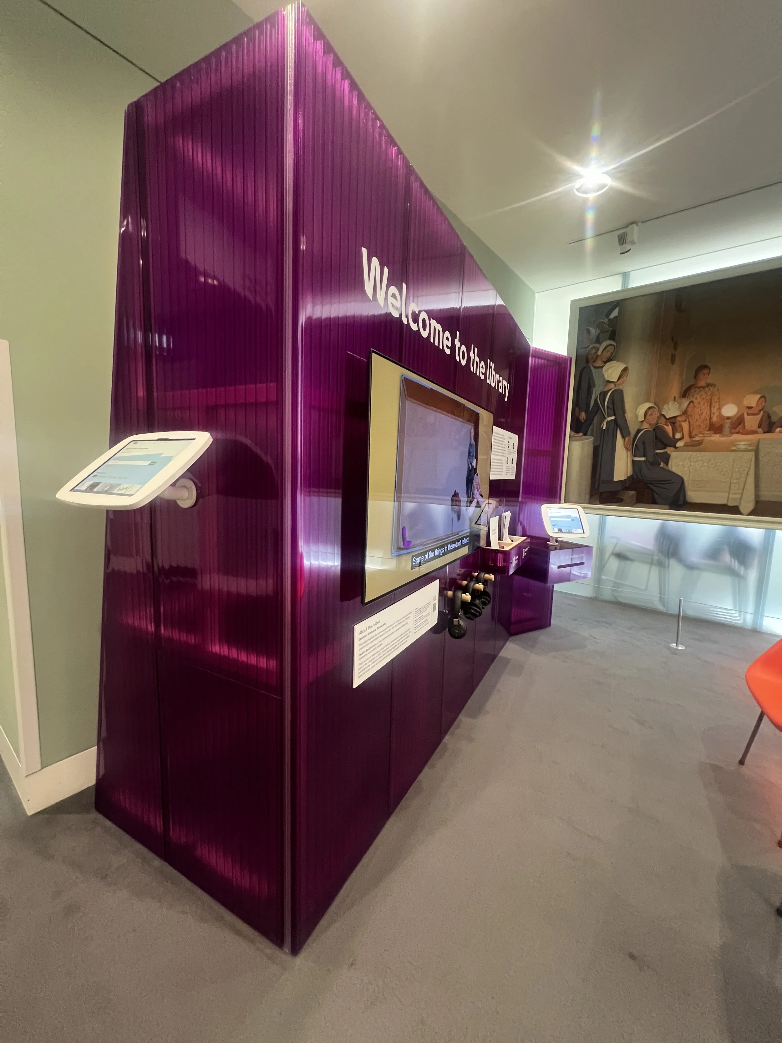 A purple interactive display at a library with a digital screen and touch controls, with a mural depicting women in traditional clothing in the background.