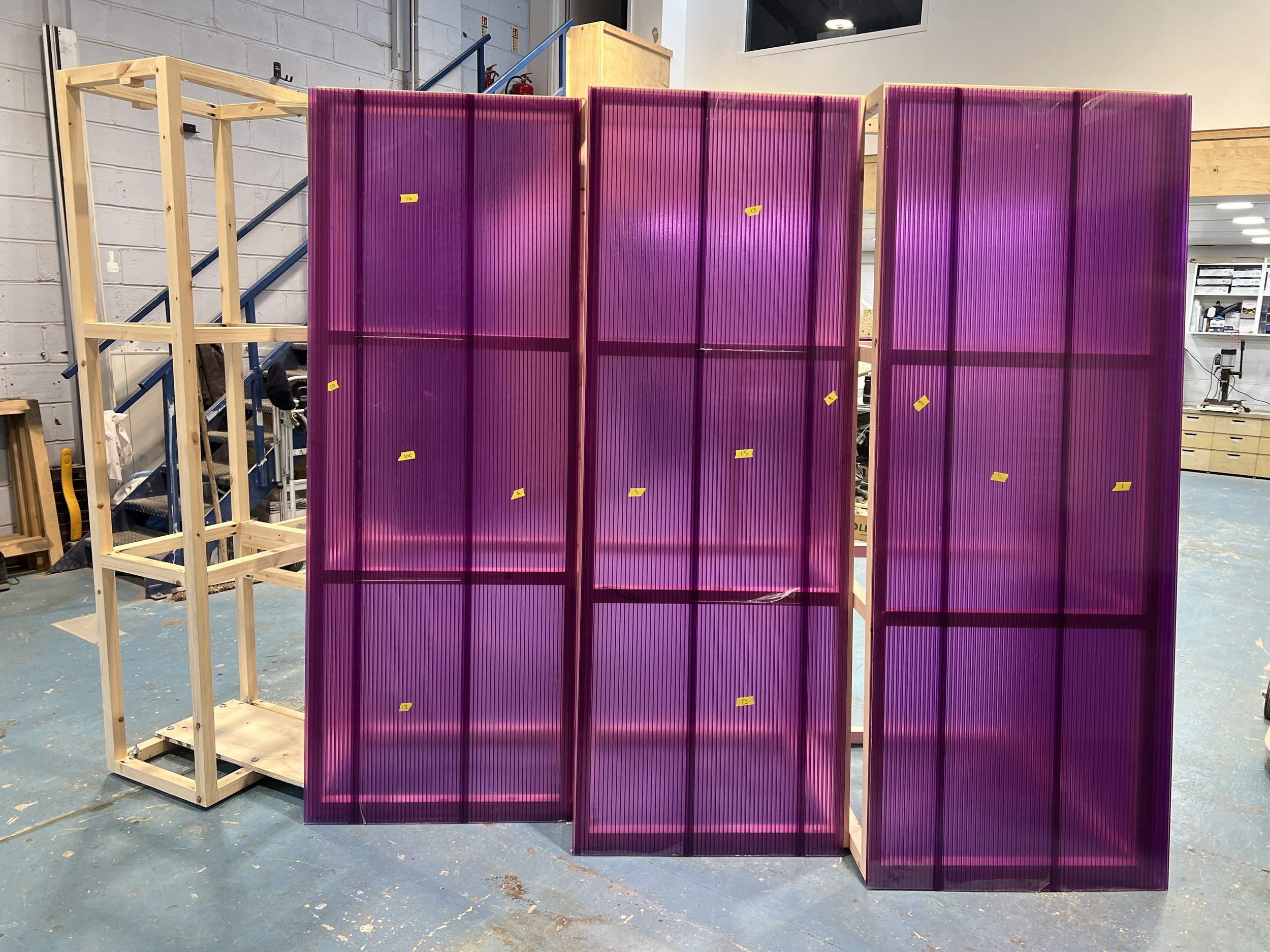 A partially assembled wooden frame holding purple plastic panels with small yellow stickers in an industrial workshop.