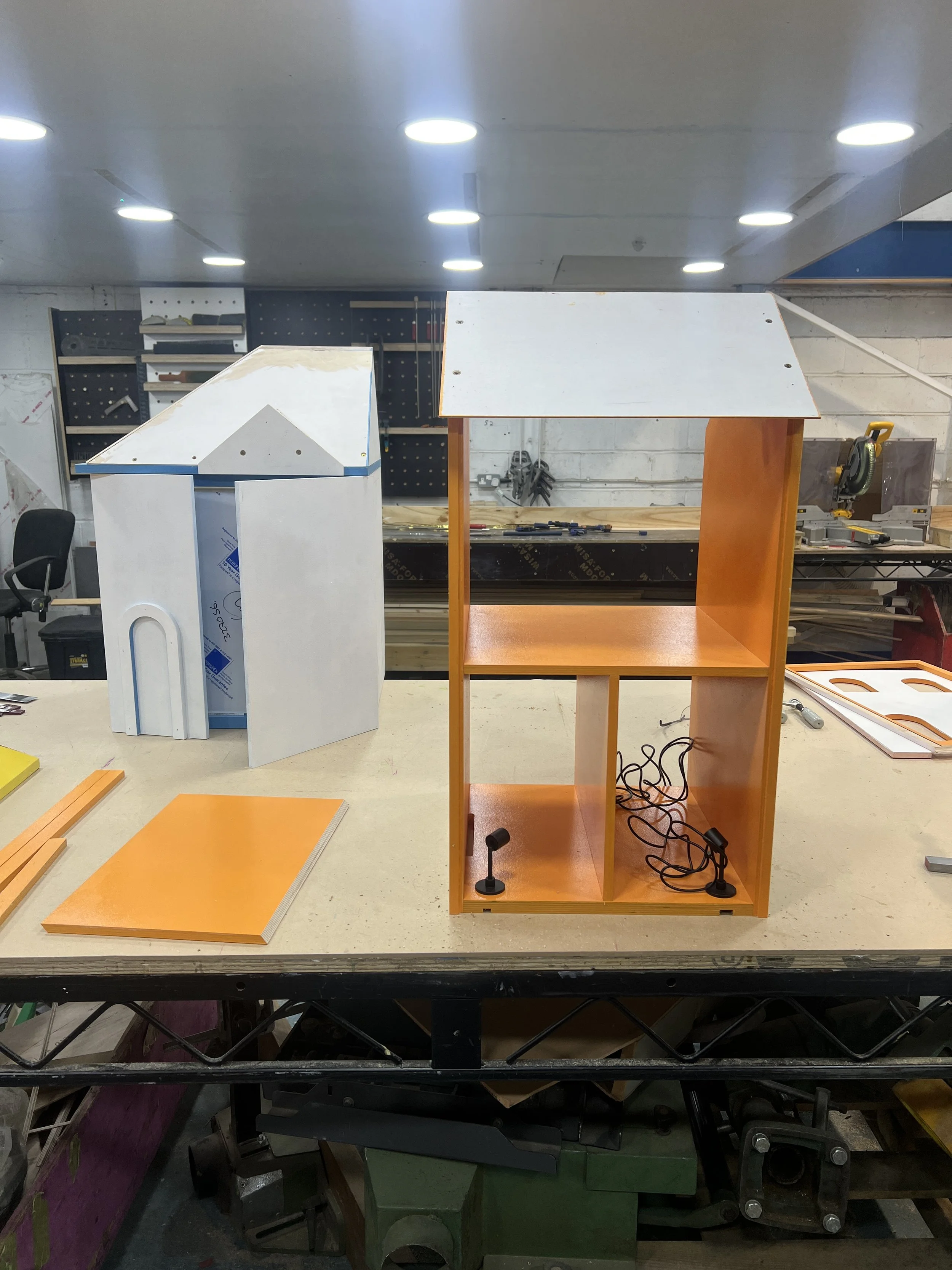 Workshop with various wooden furniture pieces, including a partially assembled orange and white bookshelf, wooden panels, and power tools on a workbench.
