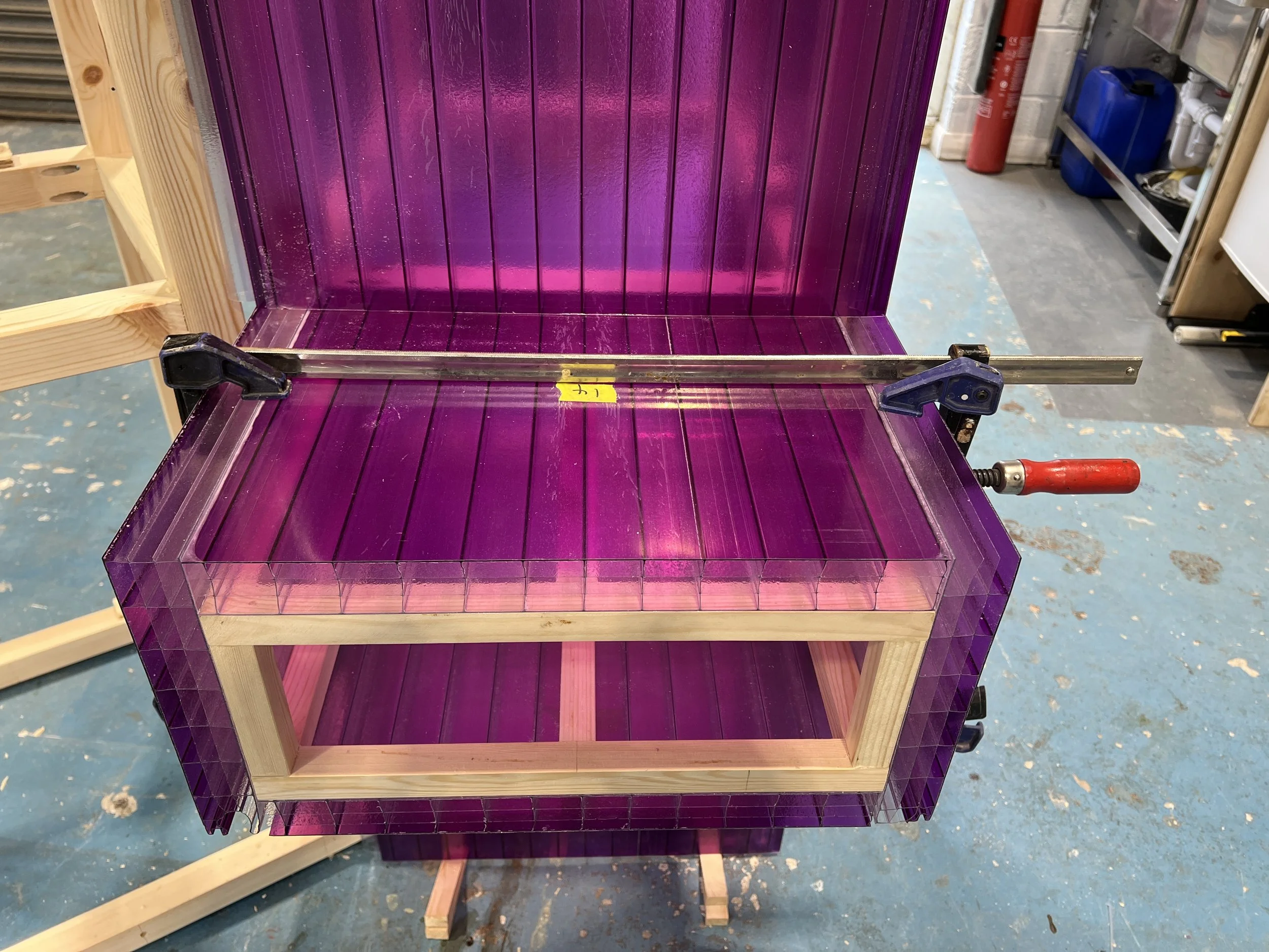 A partially assembled wooden cabinet with purple and clear acrylic panels in a workshop. Clamp tools are securing the top panel, and shelves are visible inside.