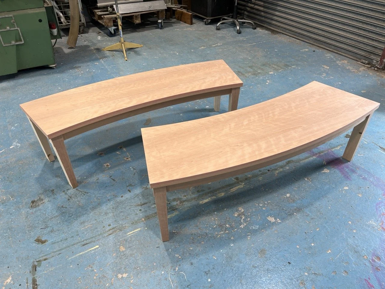 Two unfinished wooden benches with curved seats and straight legs in a workshop with a blue paint-splattered floor.