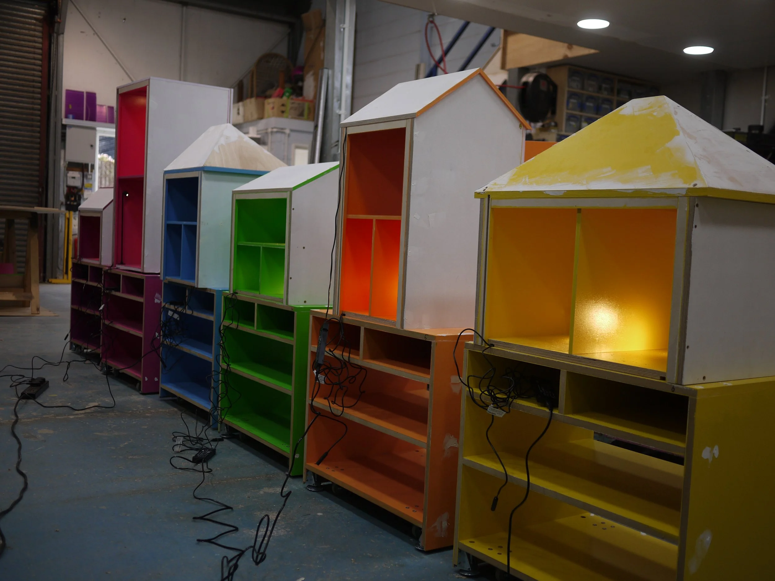 Colorful wooden shelves in a workshop, with some shelves painted in pink, blue, green, orange, and yellow, and electrical cords hanging from them.