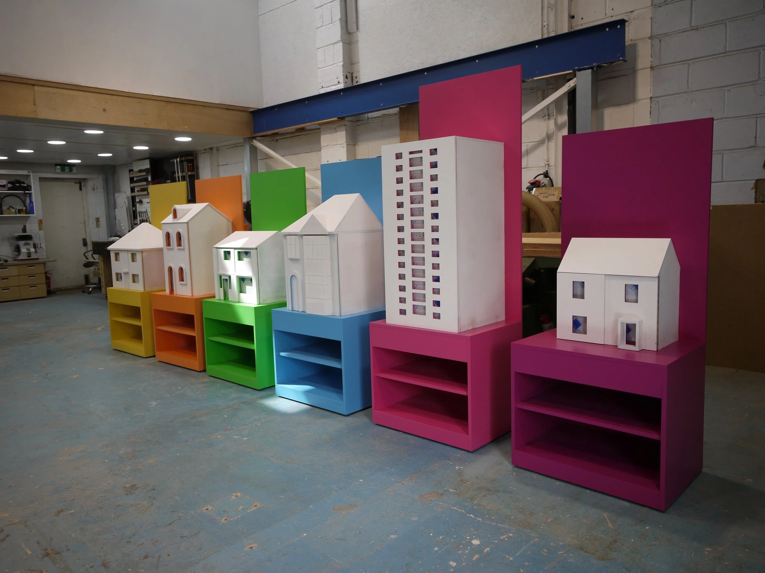 Colorful miniature building models with vibrant pink, blue, green, yellow, orange, and white colors, displayed on shelves in a workshop or art studio setting.