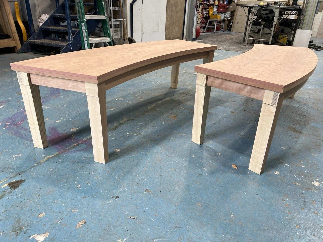Two unfinished wooden curved tables in a woodworking workshop, with visible tools and equipment in the background.