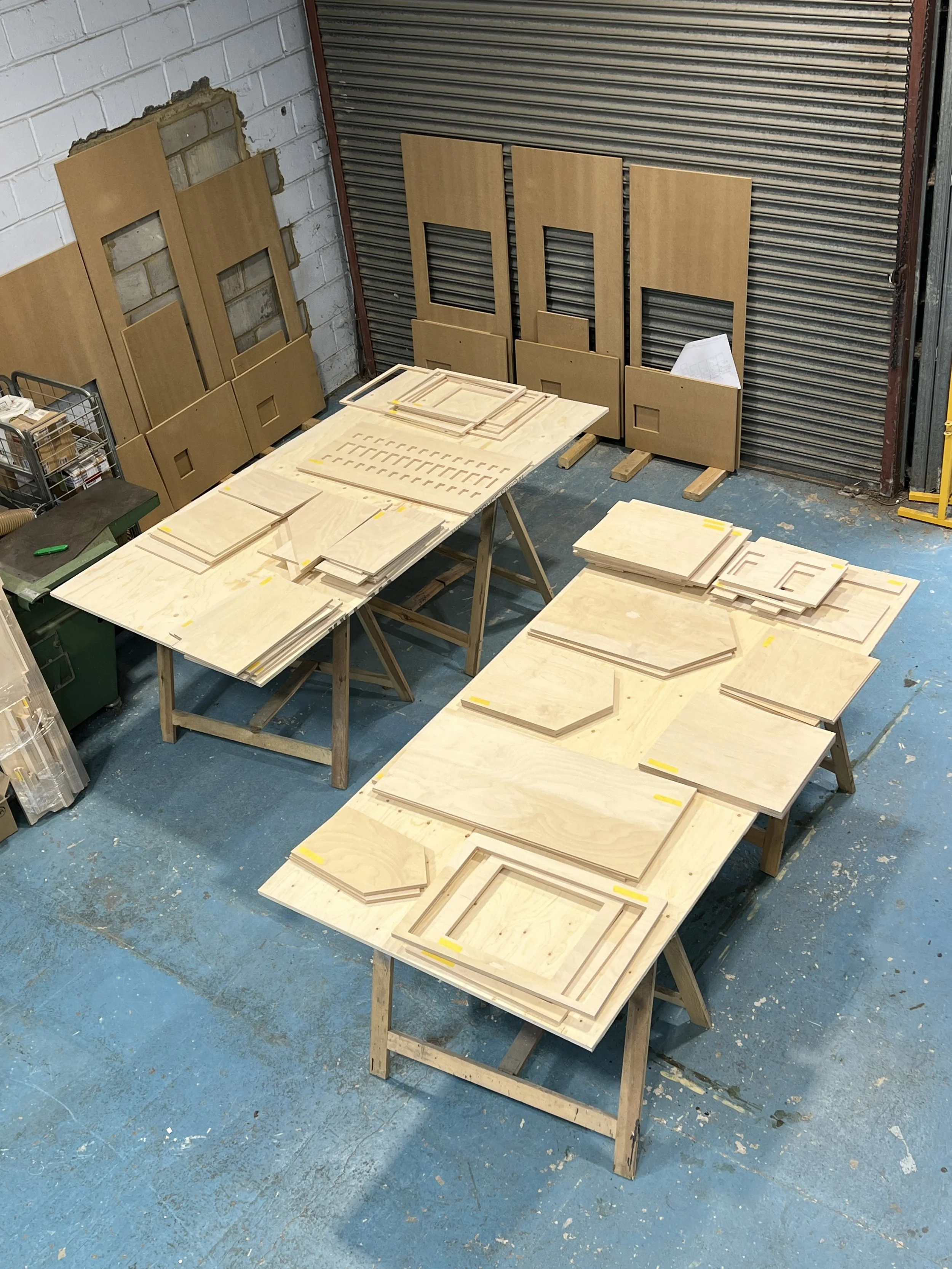 Wooden cabinet doors with cutouts laid out on tables in a woodworking shop, with unfinished walls and a roller door in the background.