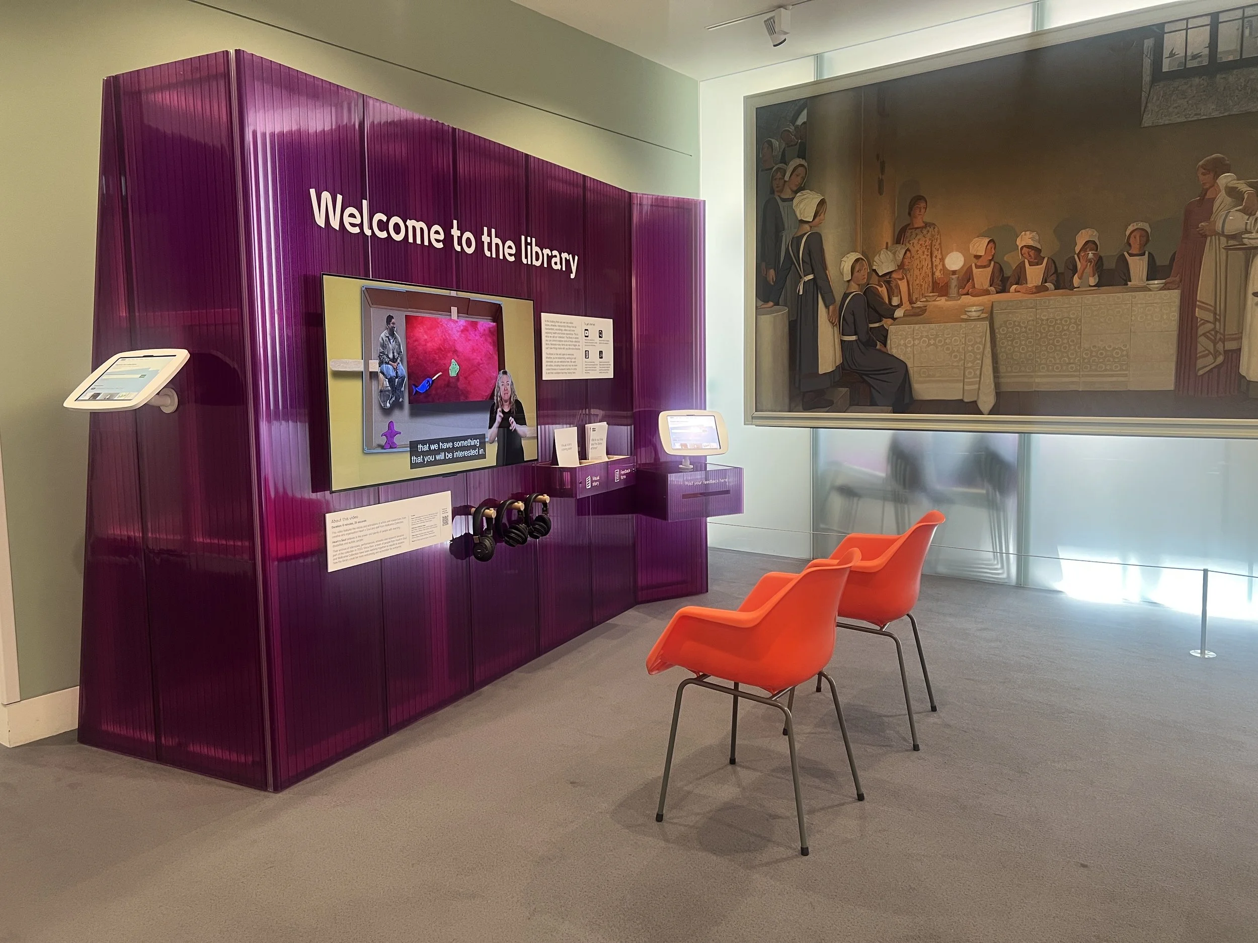 A waiting area in a library with three orange chairs, a purple information kiosk with a screen, and a large mural on the wall depicting historical women at a table. The space is well-lit with natural light.