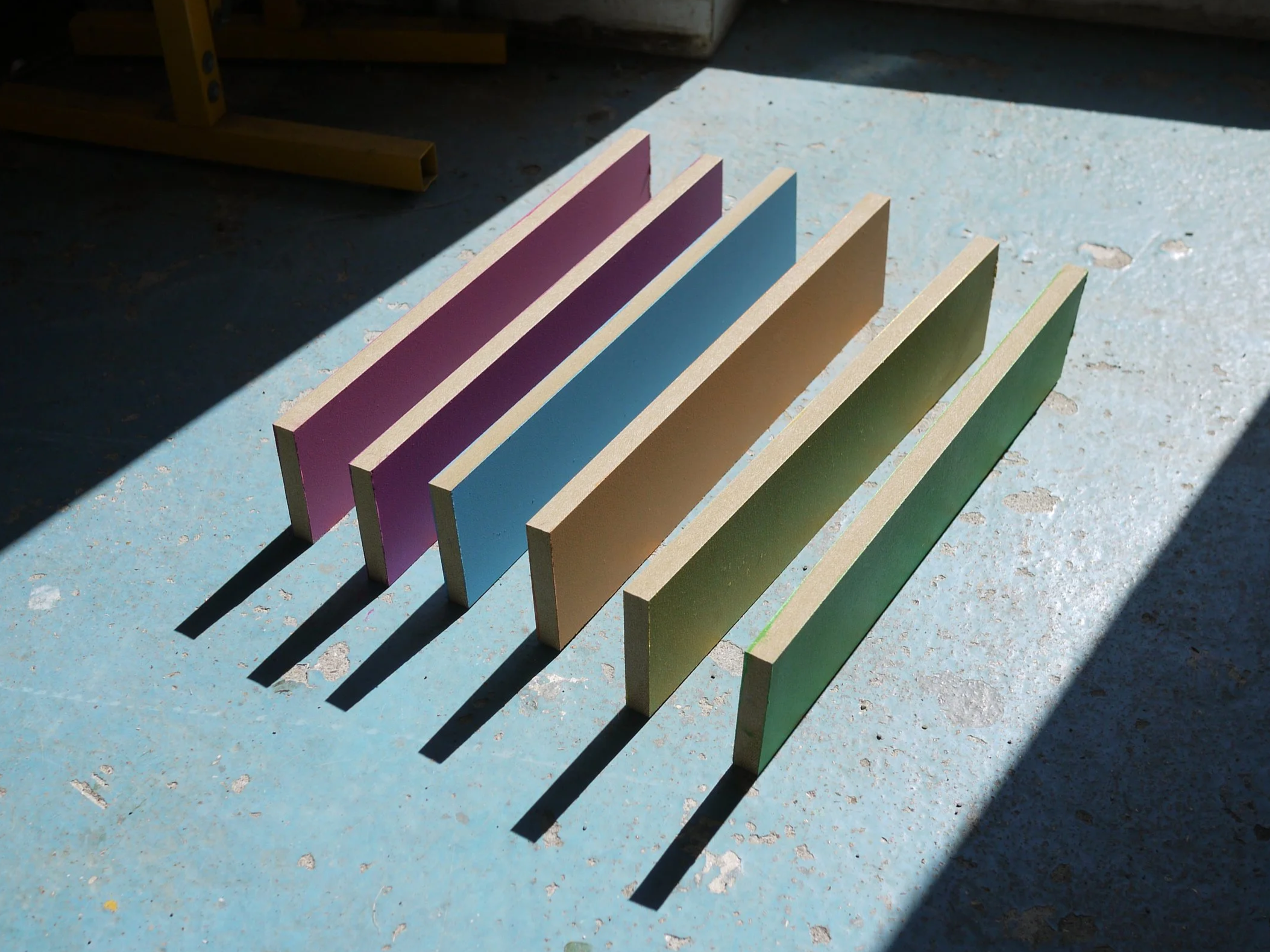 Colored rectangular tiles arranged in a fan shape on a sunlit, blue concrete surface, casting shadows.