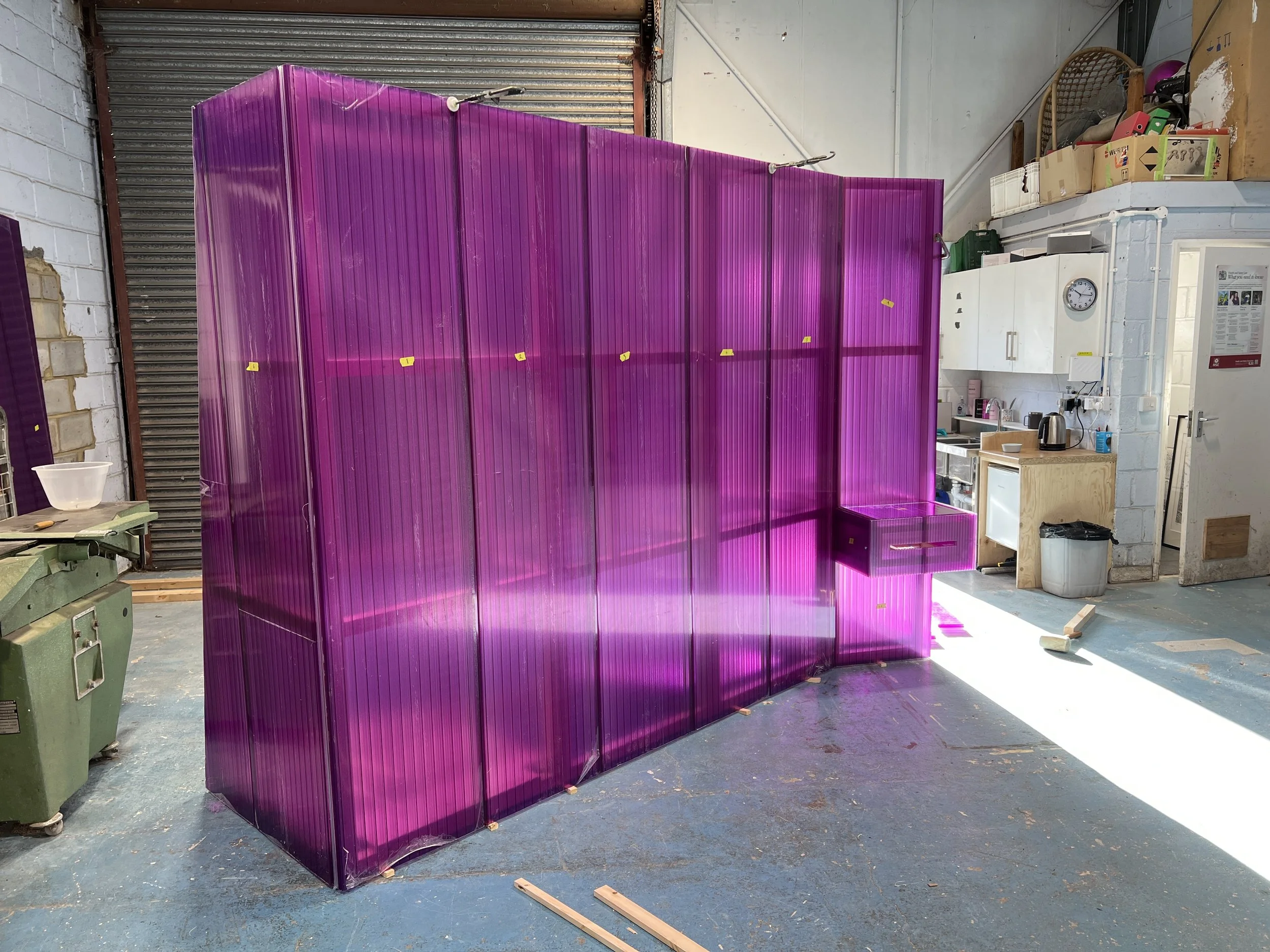 A large indoor space with a purple plastic or acrylic structure in the center, possibly a work in progress. The space has a concrete floor, a white brick wall on the left, a garage-style metal door, and a kitchenette area with a white cabinet, clock, and some kitchen appliances. Various boxes and tools are visible around the space.