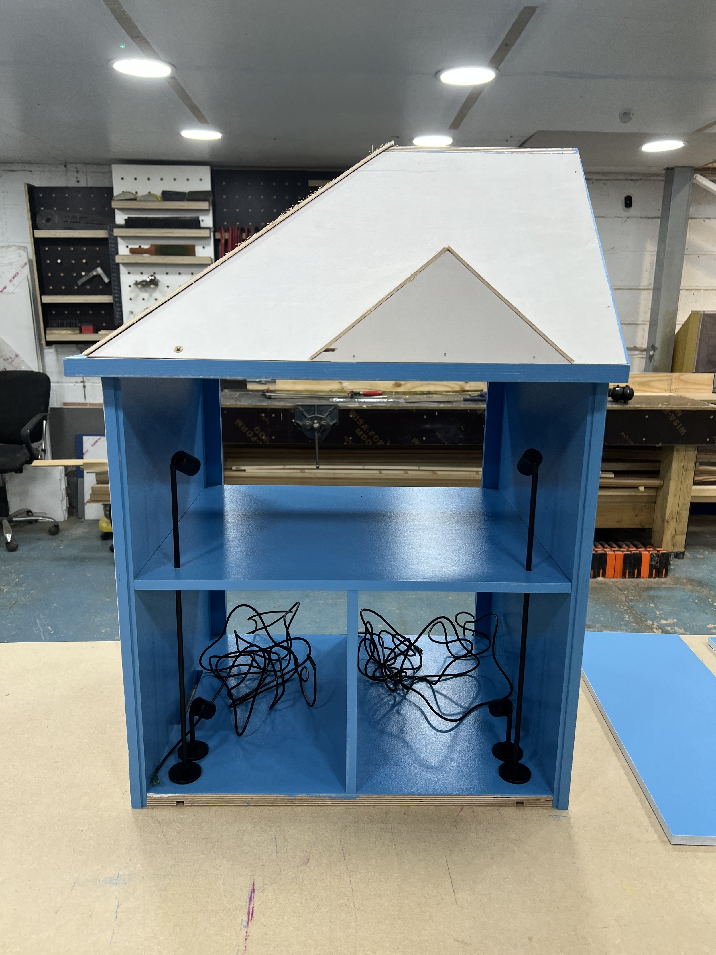 Unfinished blue wooden structure resembling a small house or booth, with wires hanging inside, set in a workshop or woodworking space.