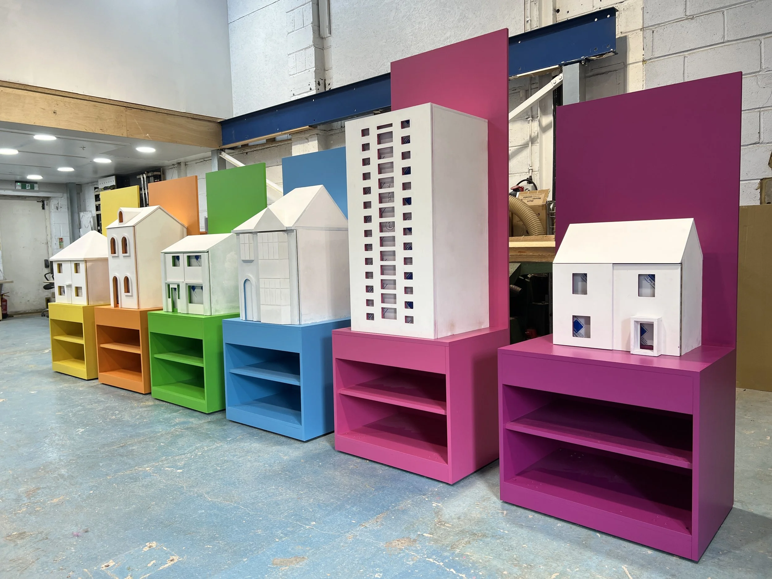 Colorful display shelves in a workshop, arranged in rows with miniature models of houses on top, against a white brick wall, with brightly colored back panels in pink, blue, green, yellow, and orange.