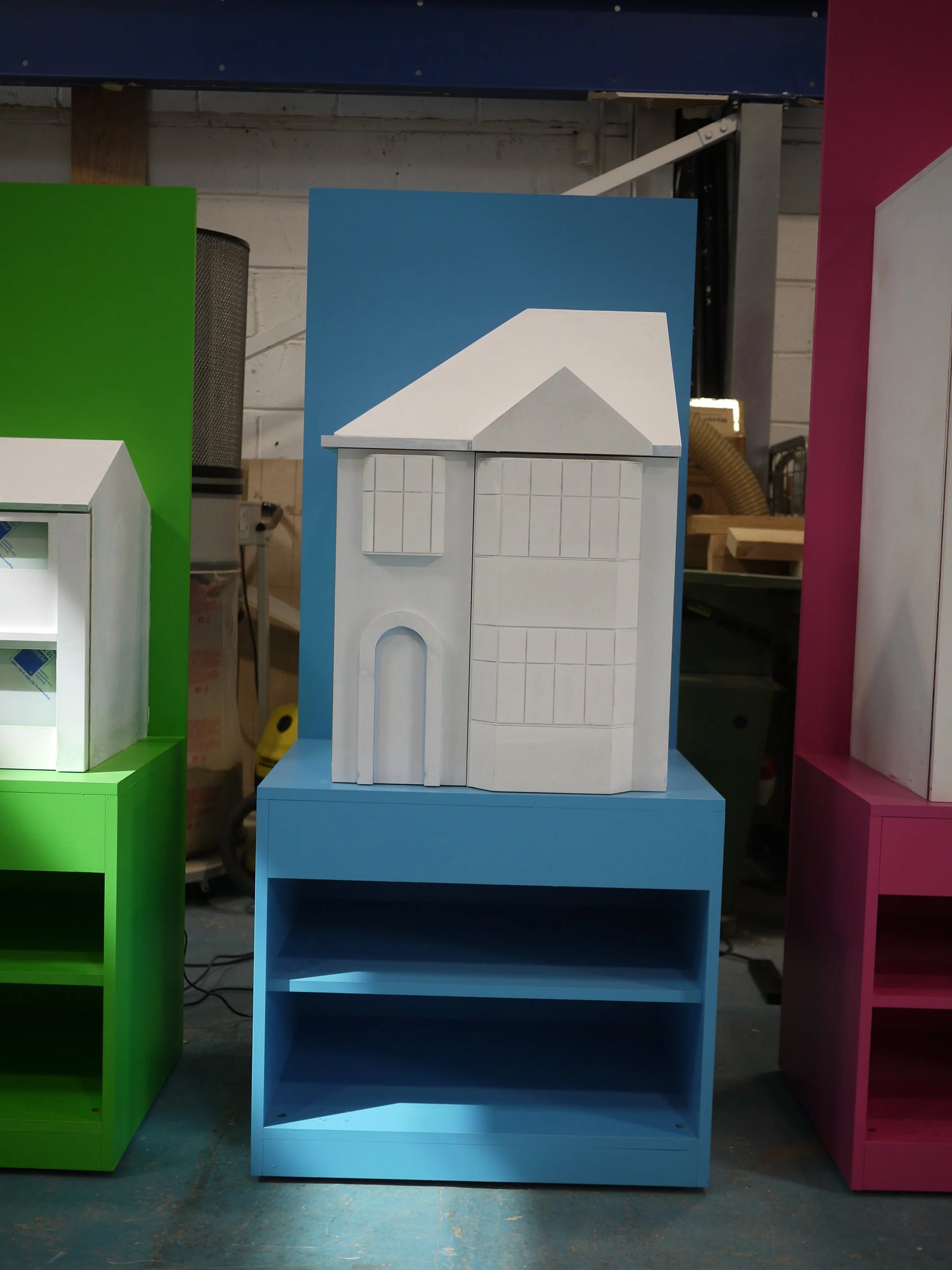 Colorful wooden display shelves in green, blue, and pink, with the blue shelf holding a white model house with a gray roof.