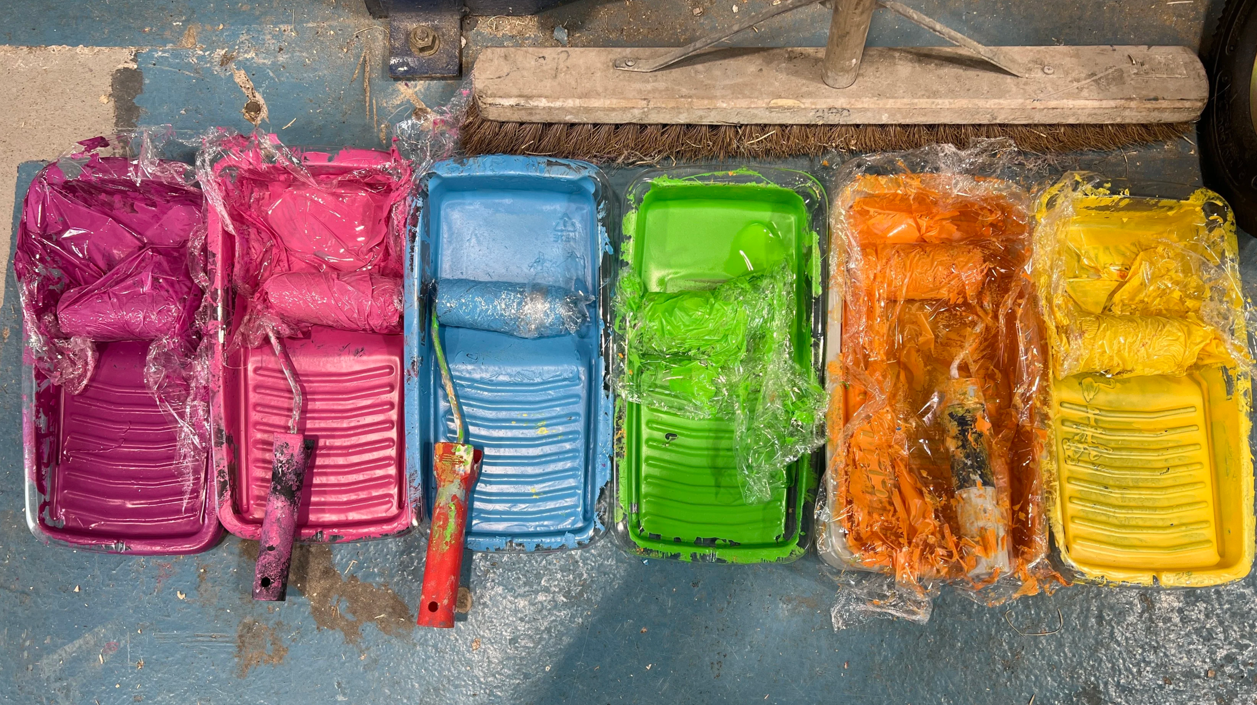 Surfaces of plastic paint trays in pink, blue, green, orange, and yellow with paint remnants and paintbrushes, placed on a concrete floor with a dustpan and broom above.