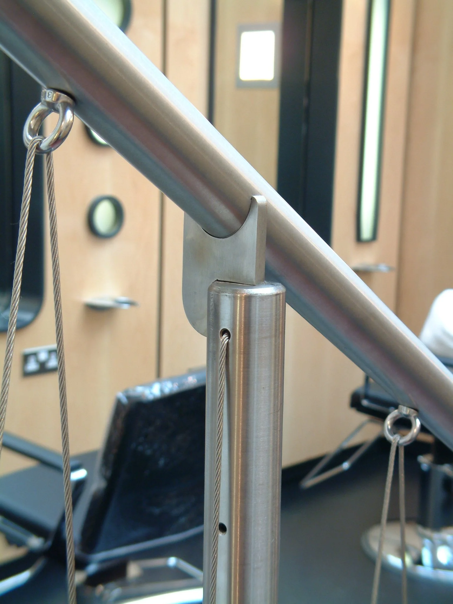 Close-up of a stainless steel handrail on a staircase, with a chain and loop on the side, and blurred background showing chairs, tables, a wall with a window, and an electrical outlet.